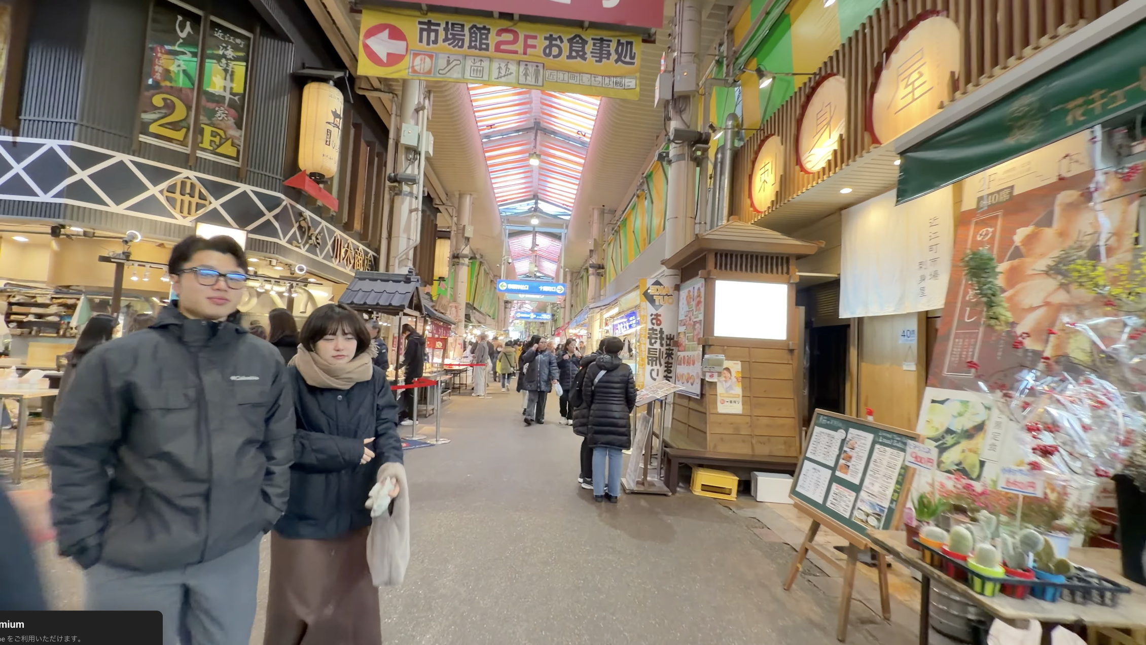 市場館2階お食事処の案内看板の下を歩くカップル、奥に近江町市場のアーケード