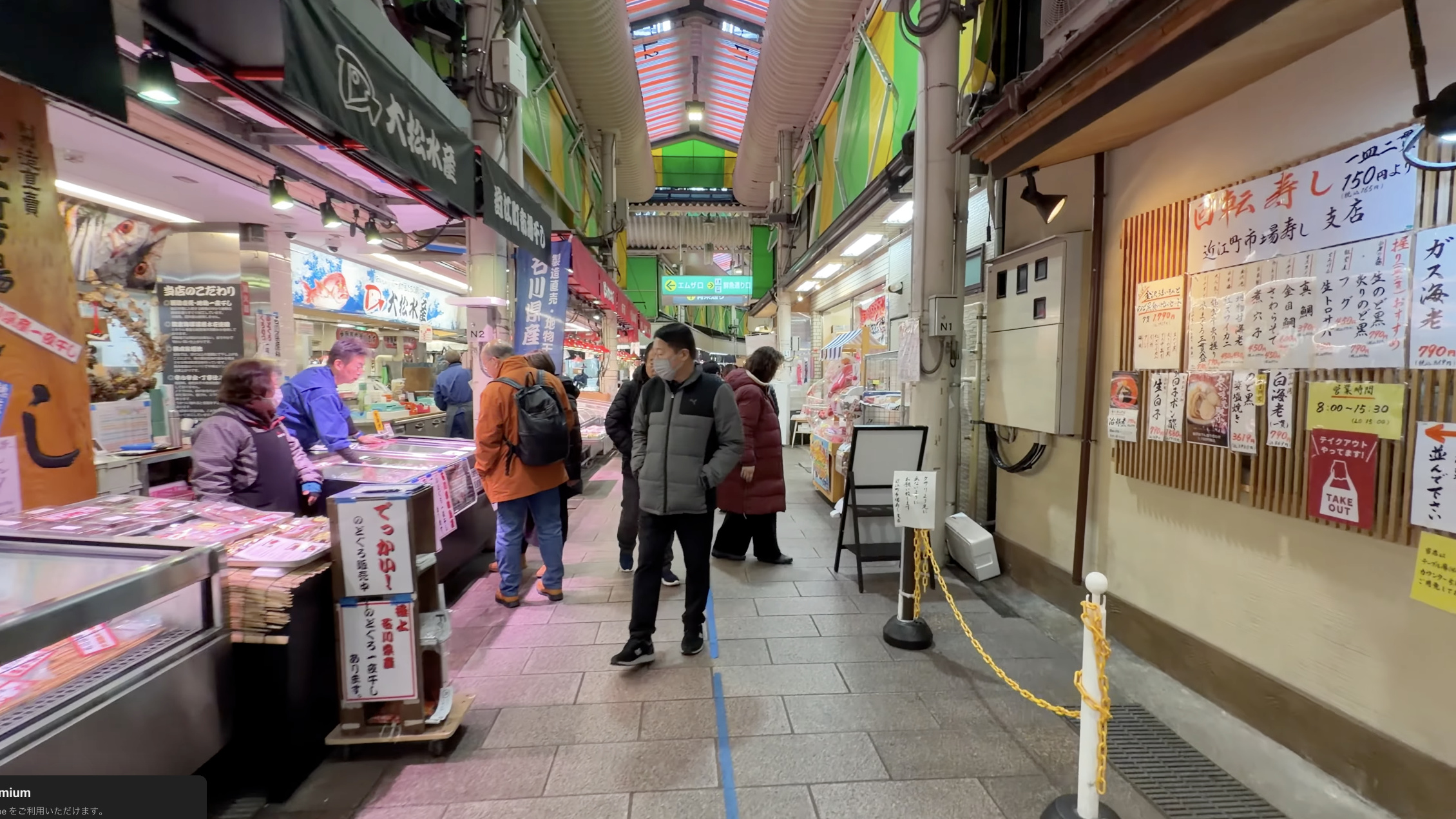鮮魚店「大松水産」の看板とショーケース、隣に「近江町市場寿し支店」の回転寿司メニュー、通りを歩く買い物客
