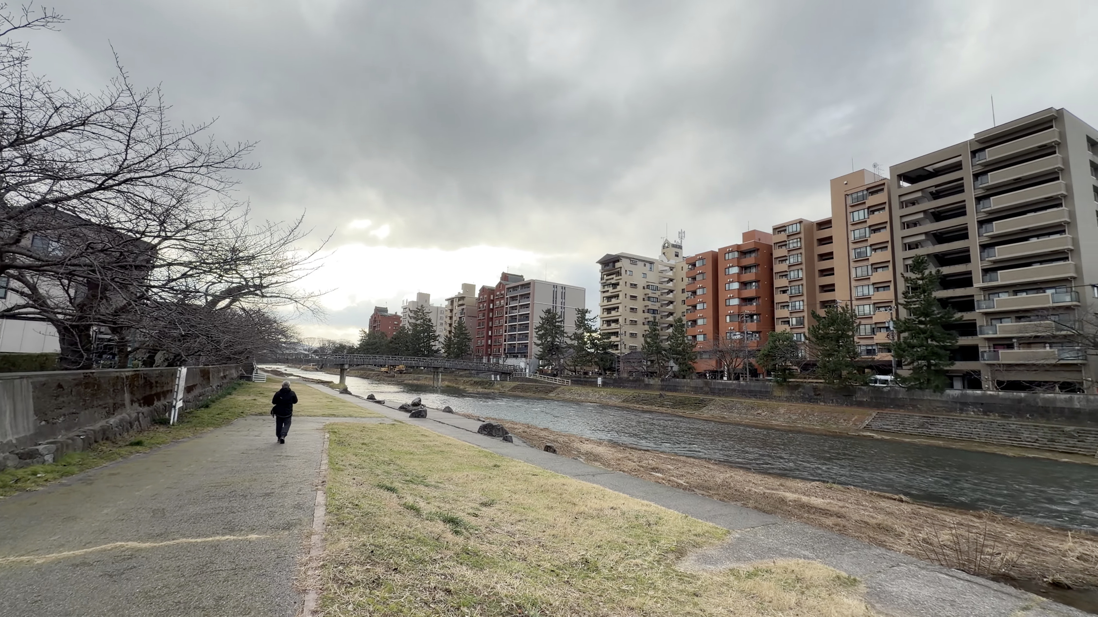 浅野川の流れと川沿いの散策路、後ろ姿で歩く人、対岸のビル群