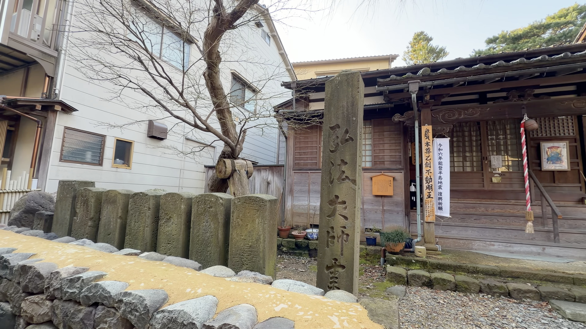 木造の寺院、弘法大師堂と刻まれた石碑、本堂脇に「令和六年能登半島地震 復興祈願」の幟