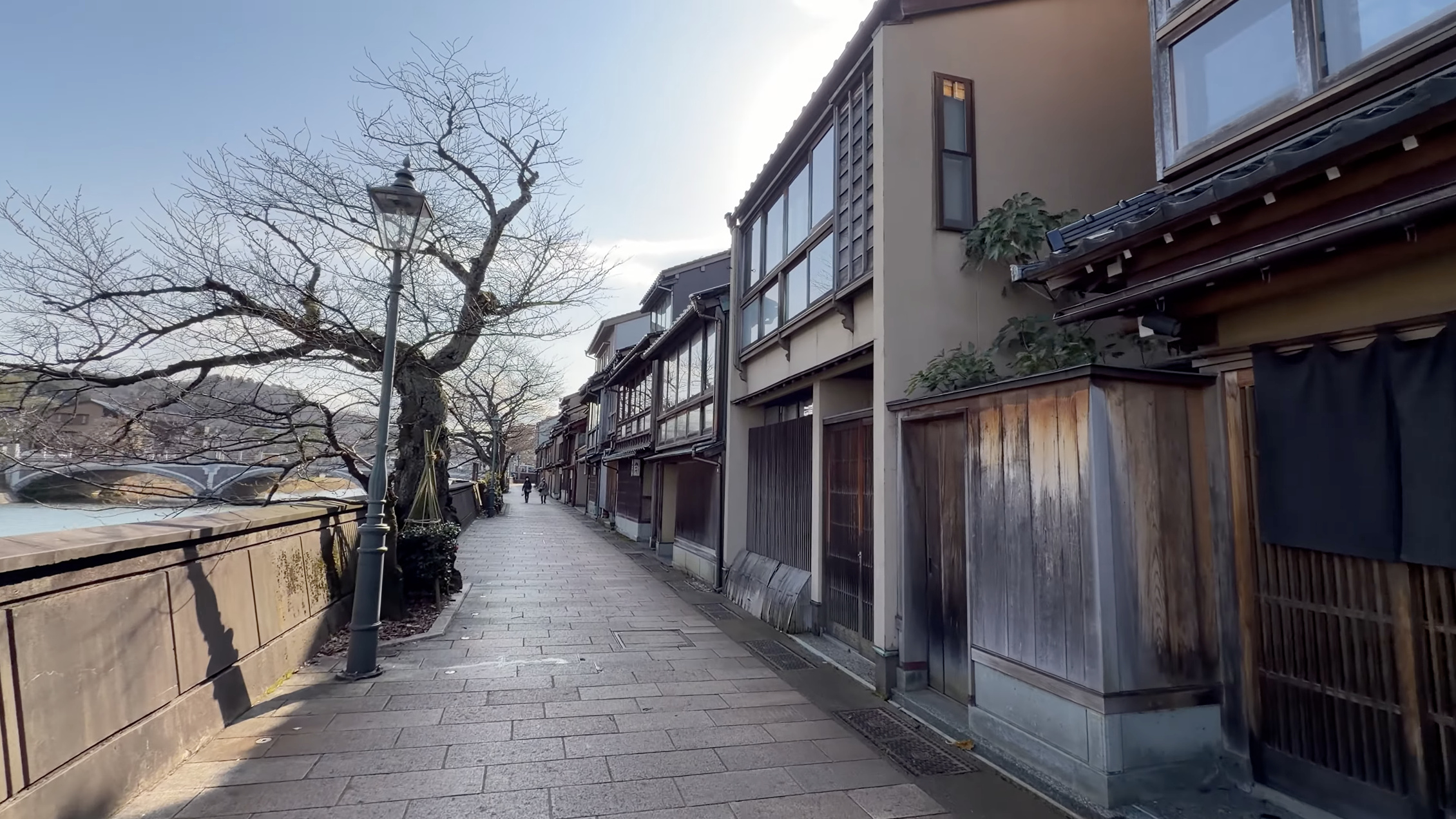 浅野川沿いの主計町の通り、古い街灯と葉の落ちた桜並木、奥に橋、川沿いを歩く人影