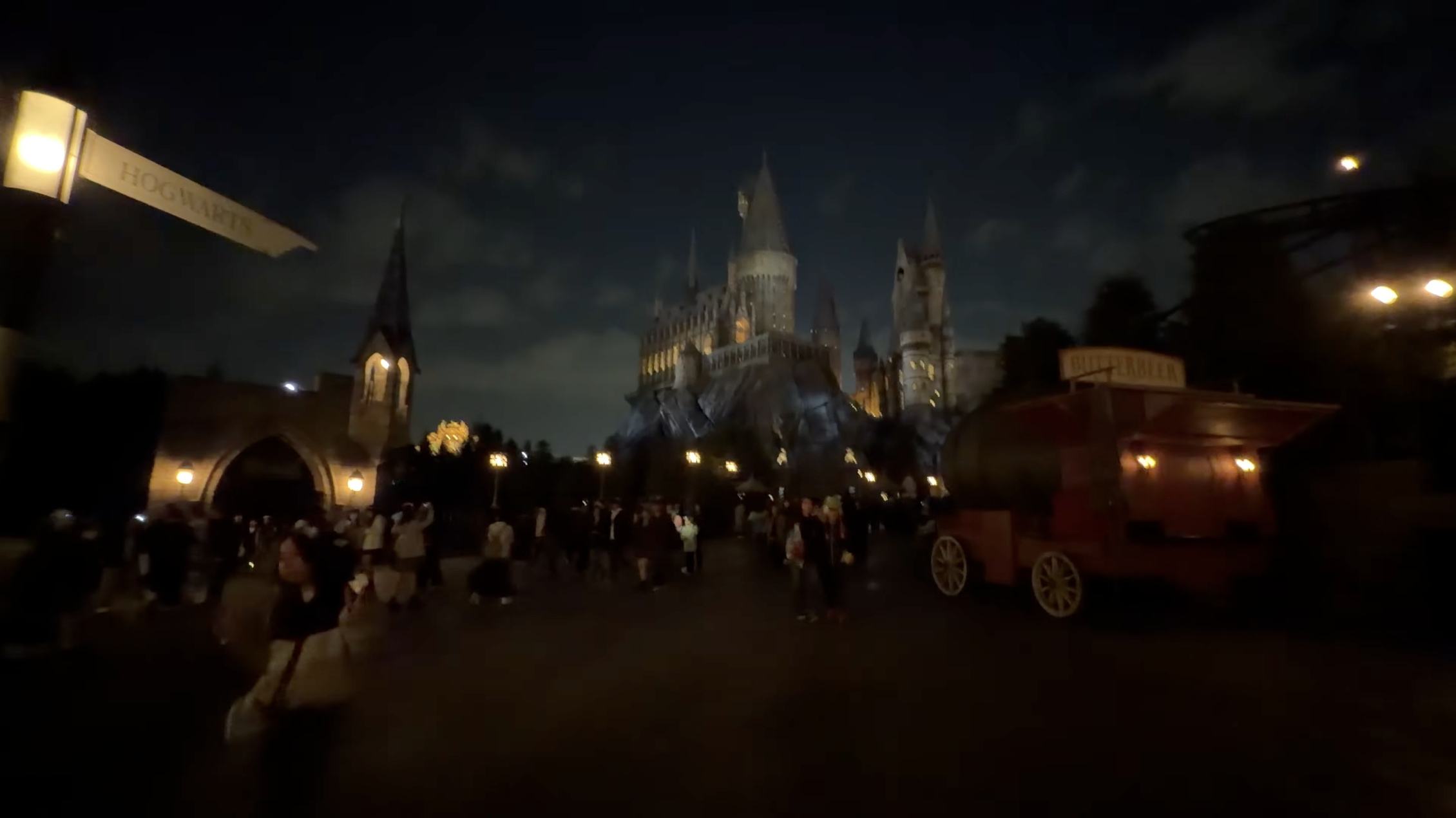 「HOGWARTS」と書かれた道標、その先に夜空に浮かぶホグワーツ城、右手にバタービアの屋台