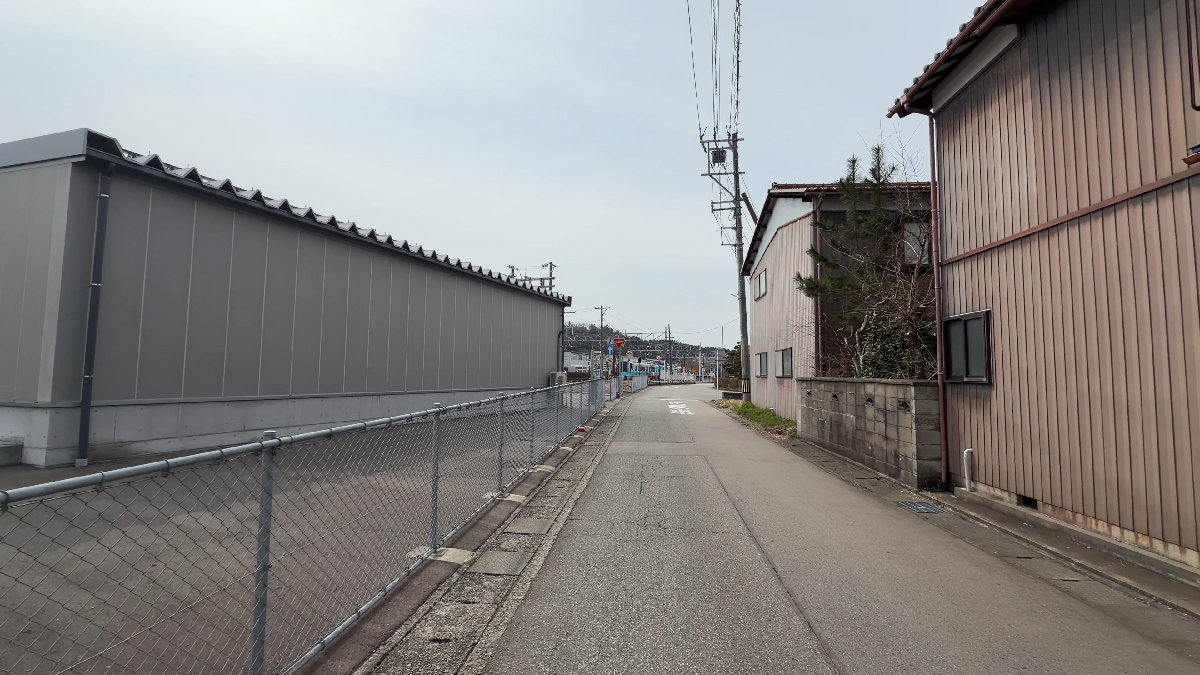 線路沿いの細い路地、灰色の倉庫の壁とピンク色の住宅、奥にIRいしかわ鉄道の車両