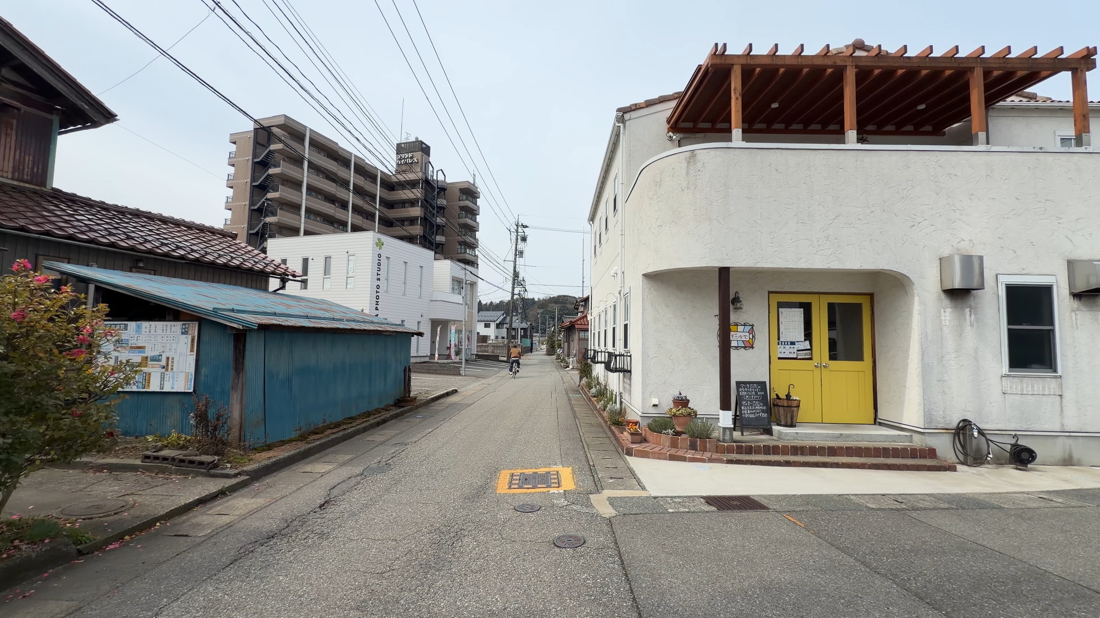 白壁の建物の角に構える黄色い両開きドア、木製のパーゴラのある外観
