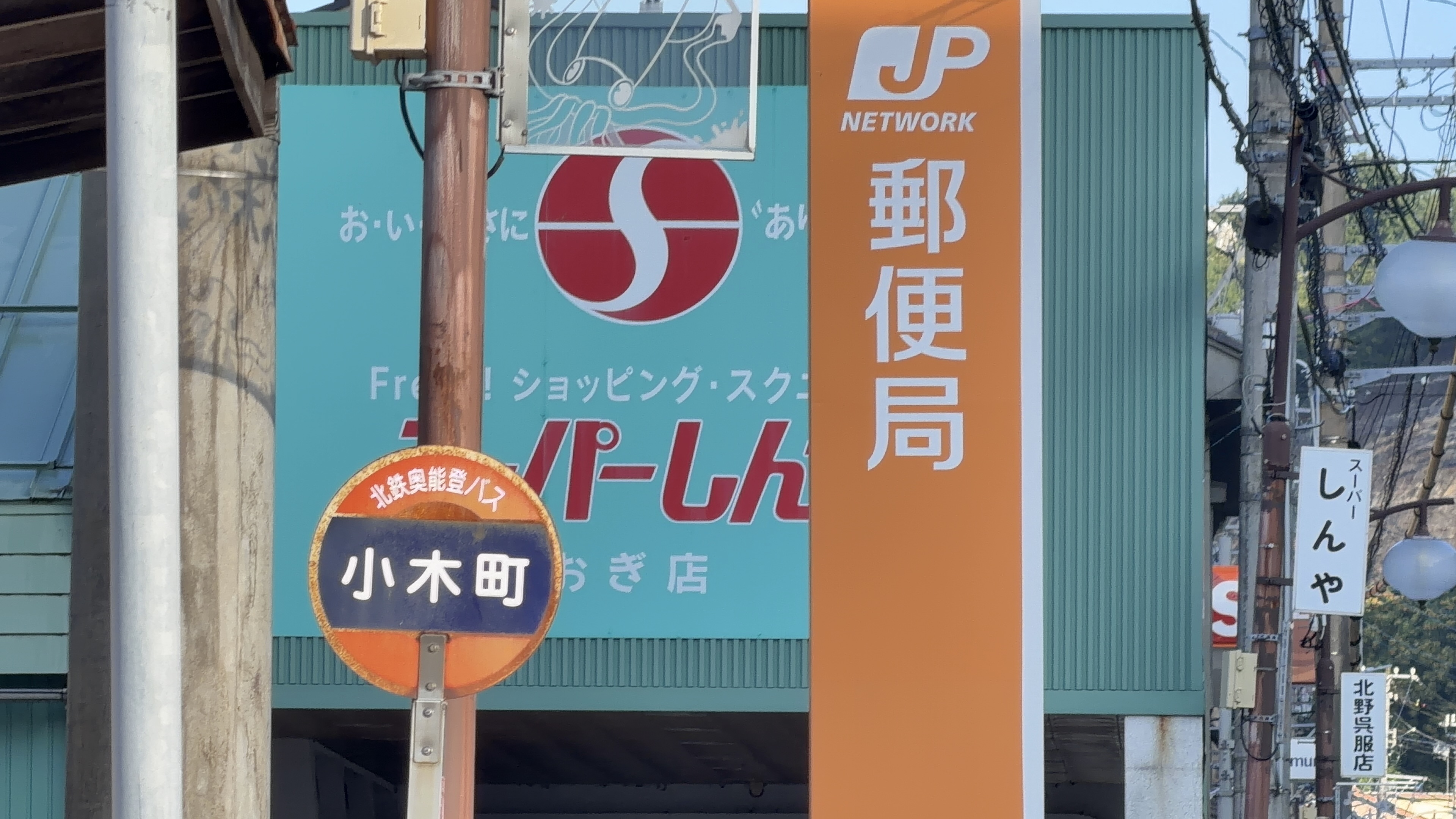 スーパーしんやの看板と郵便局、丸い小木町バス停