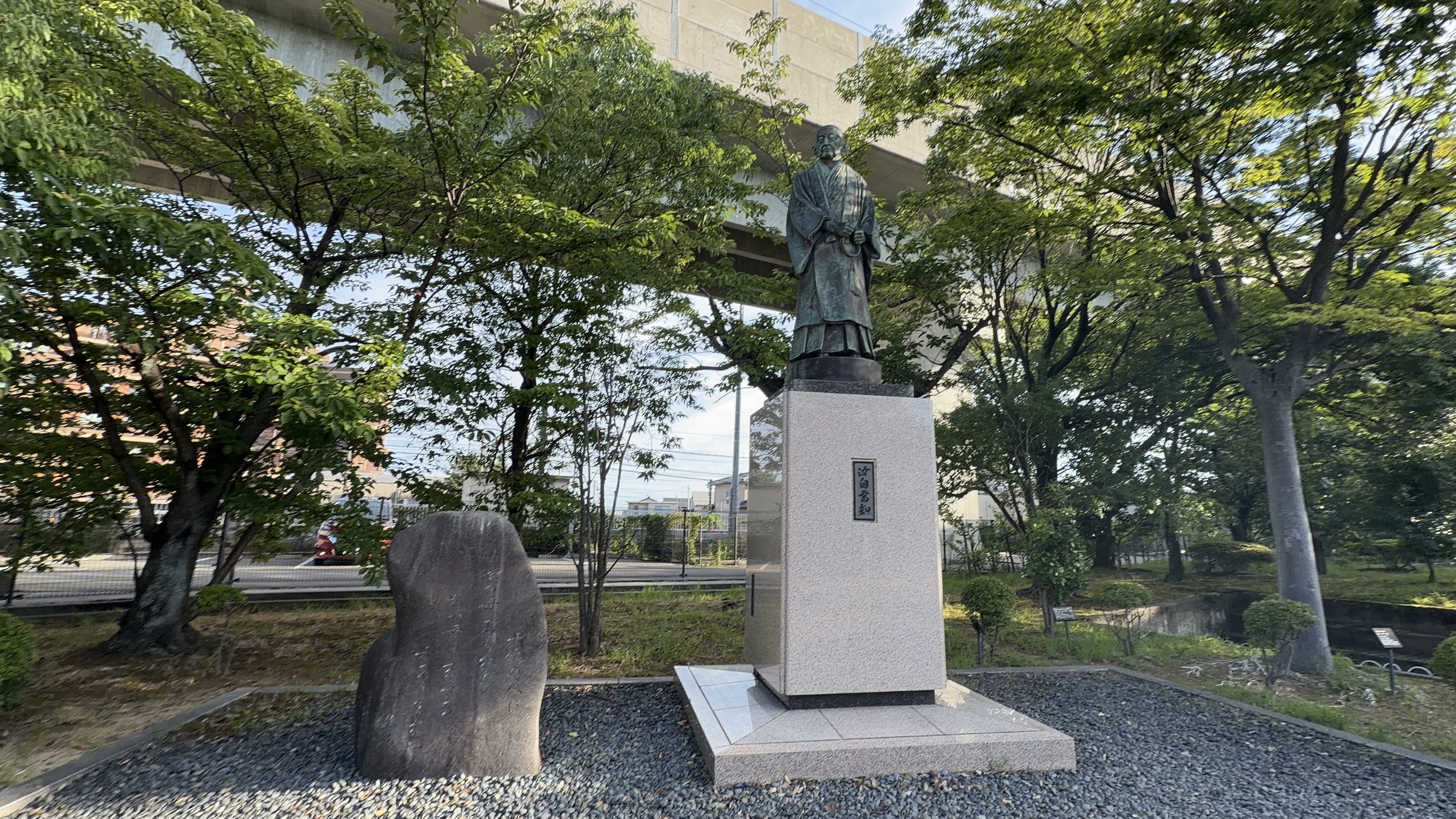 暁烏敏の銅像、羽織すがたで数珠を持つ、高架の下、隣に石碑