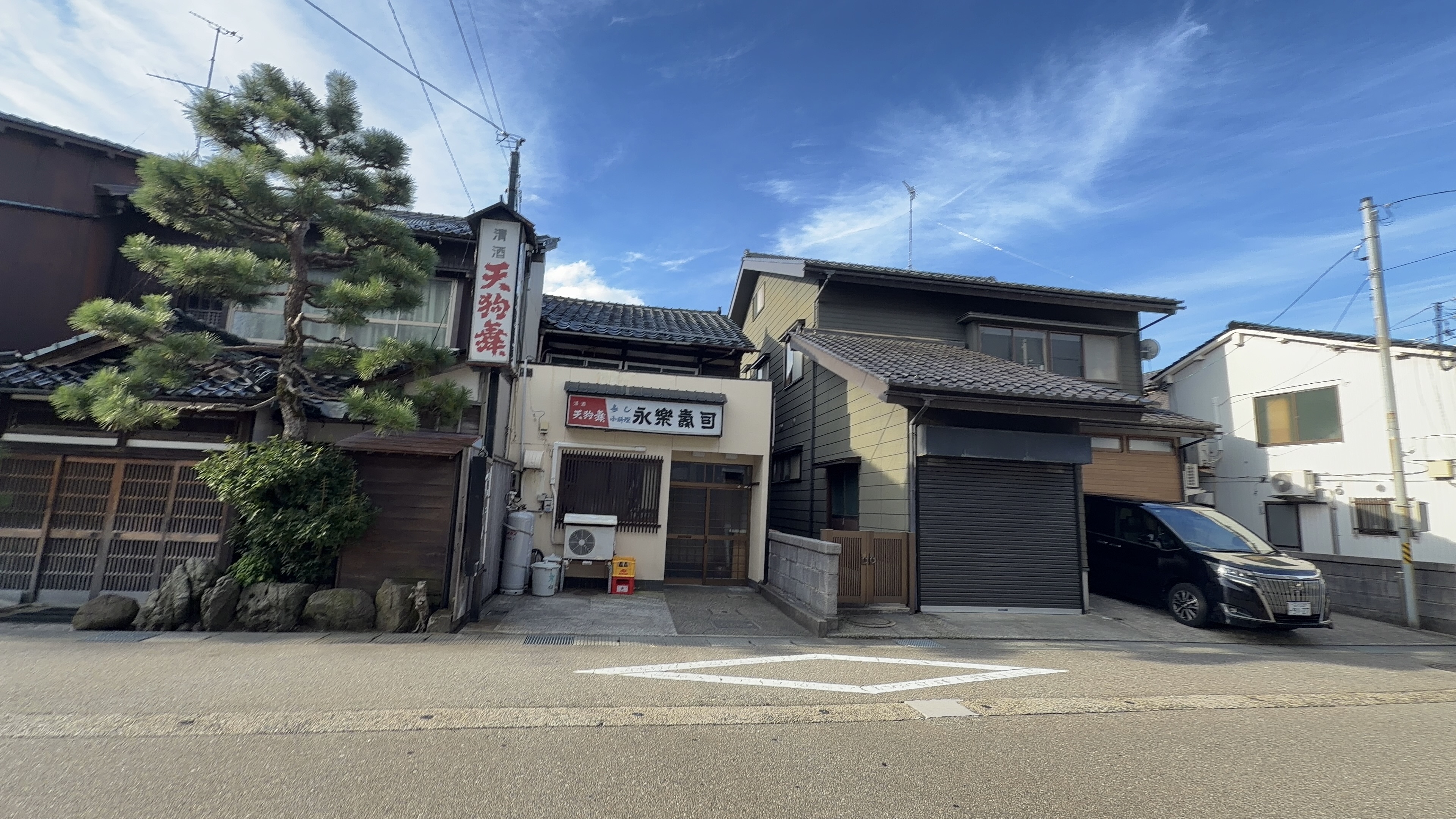 清酒「天狗舞」の看板を掲げた酒店と、隣の「永楽鮨」の店舗、松の木のある住宅街の通り