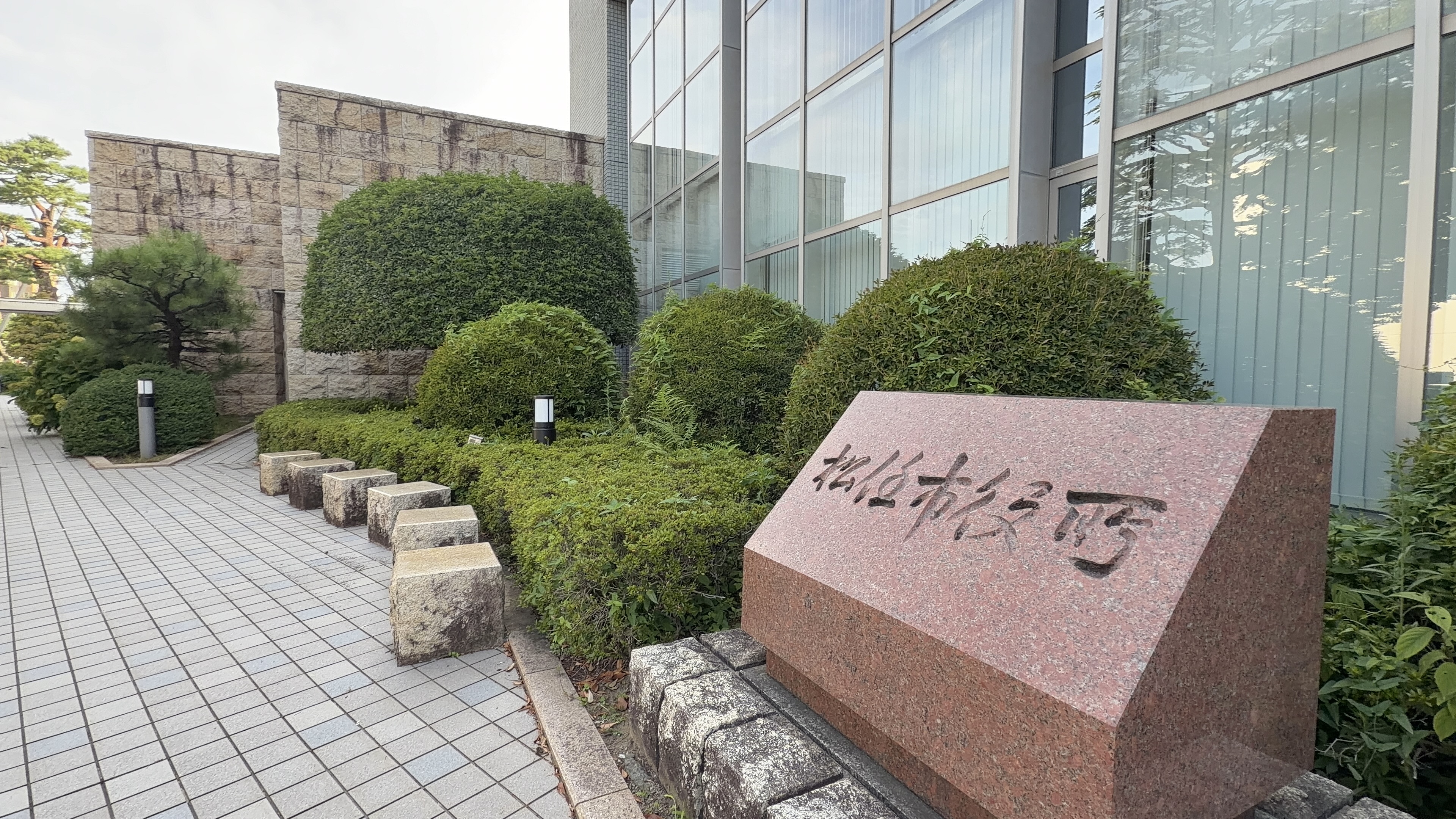 ピンク色の御影石の石碑「松任市役所」、植え込みの向こうに白いタイル張りの建物のガラス窓