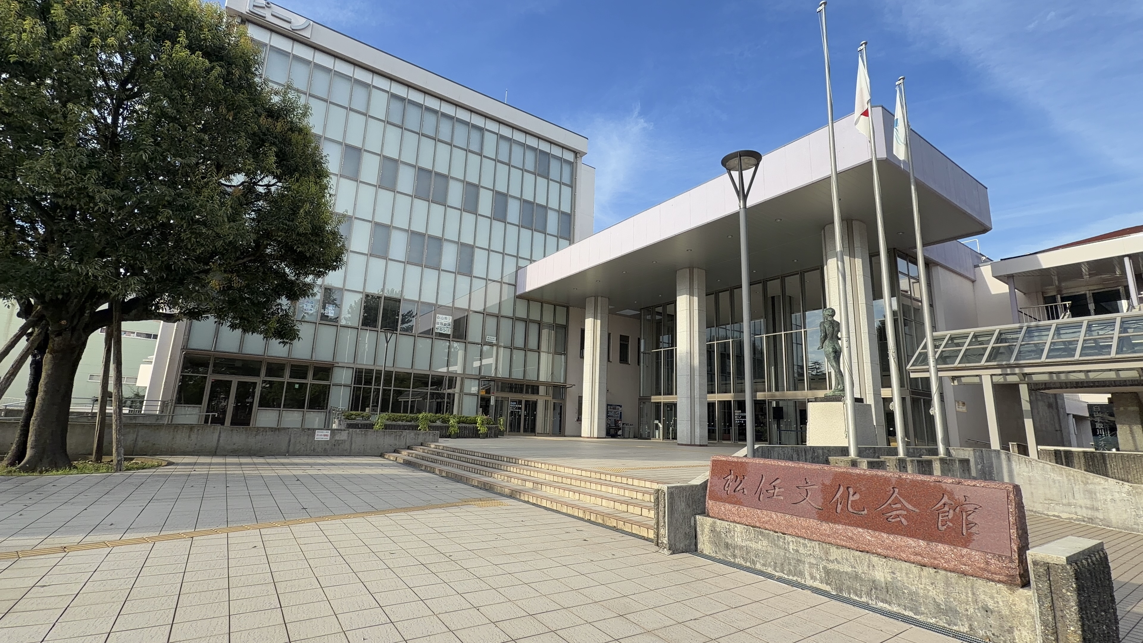 松任文化会館の建物、ガラス張りのエントランスと銅像、「松任文化会館」の石碑、連絡デッキ