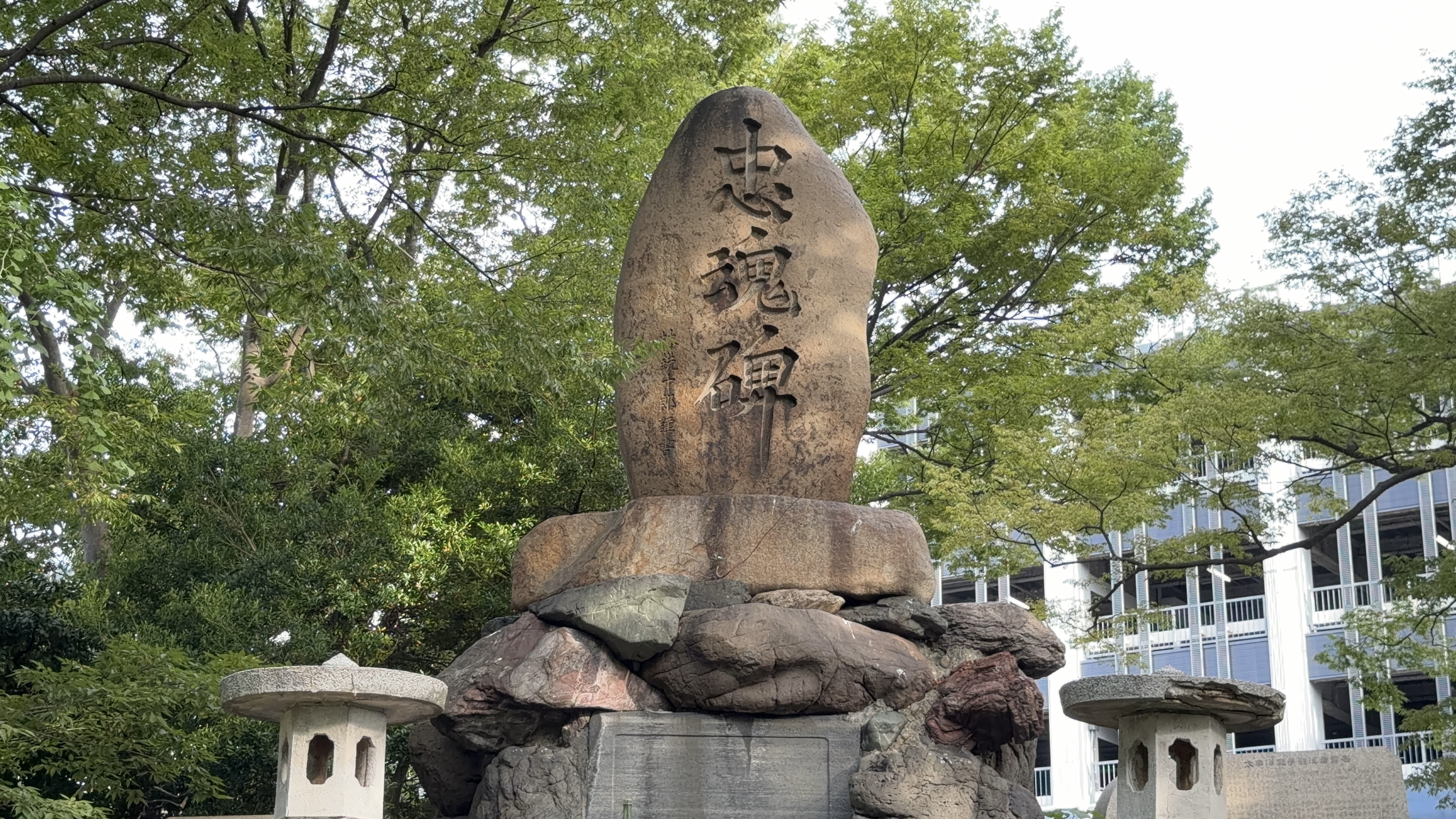 「忠魂碑」と彫られた大きな自然石のクローズアップ、緑の木立を背景に
