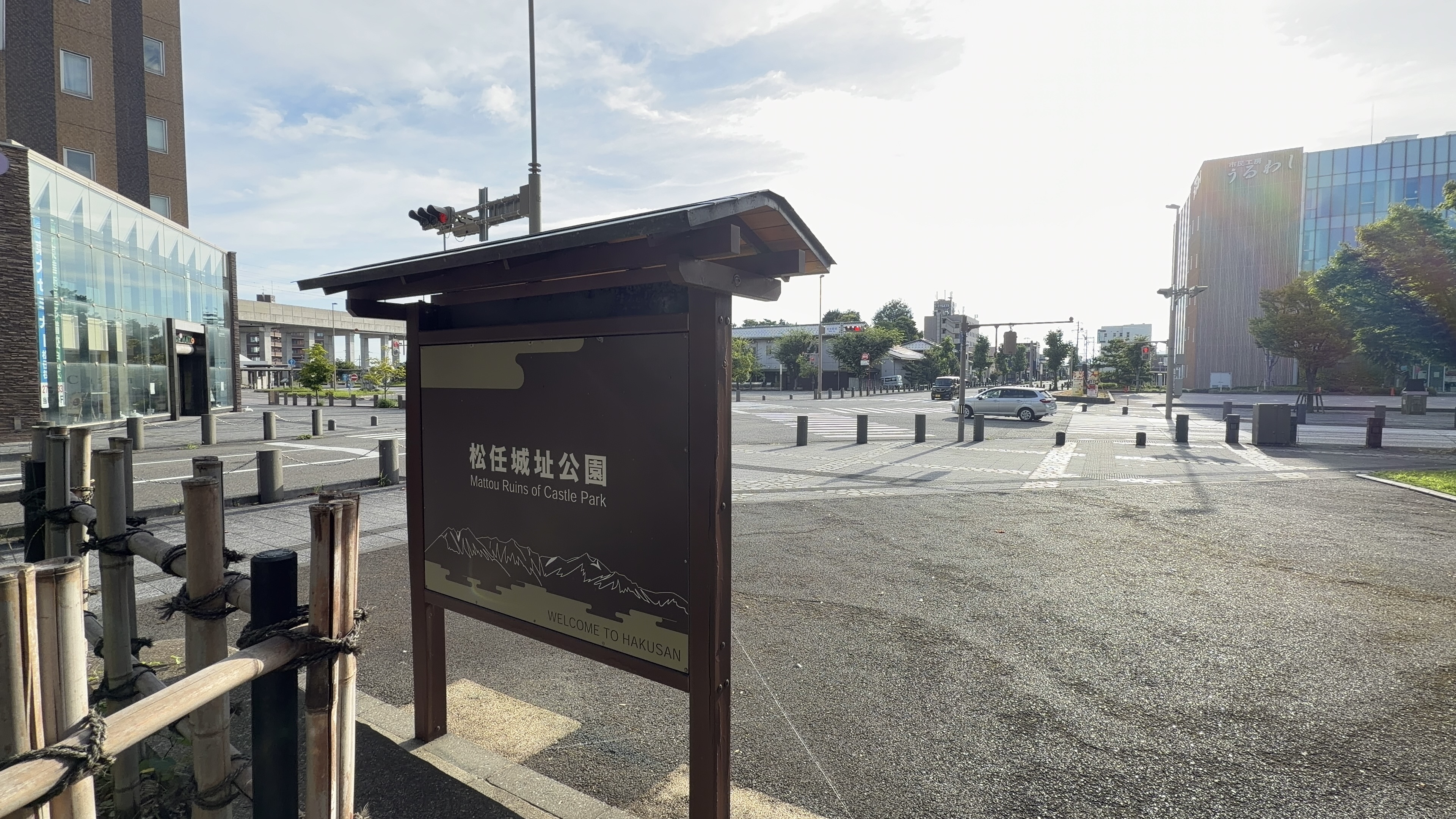 「松任城址公園 Mattou Ruins of Castle Park」の木製案内看板、白山の山なみのイラストと「WELCOME TO HAKUSAN」の文字、駅前交差点