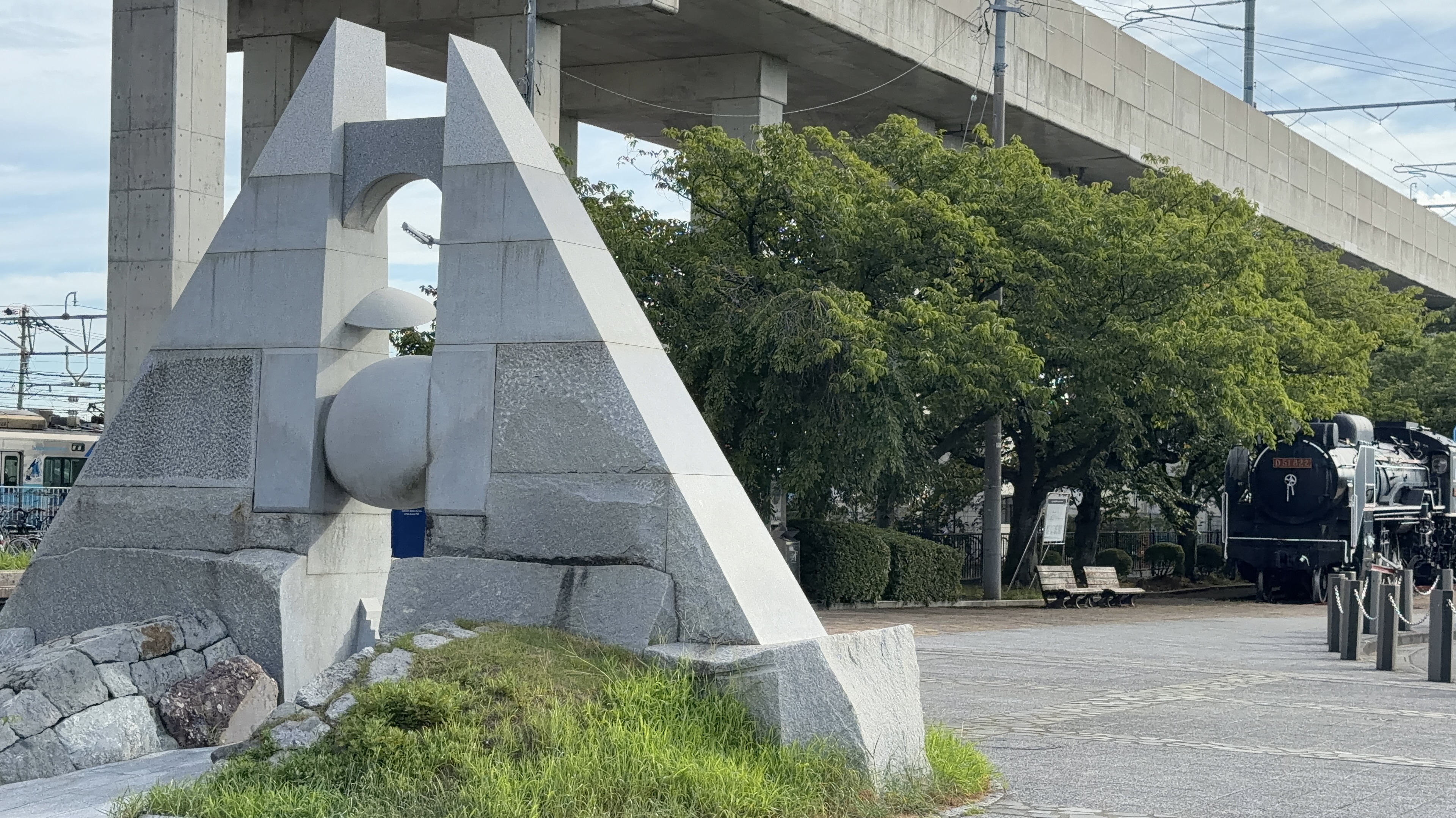 三角形の石のモニュメント越しに見える黒い蒸気機関車D51、北陸新幹線の高架下