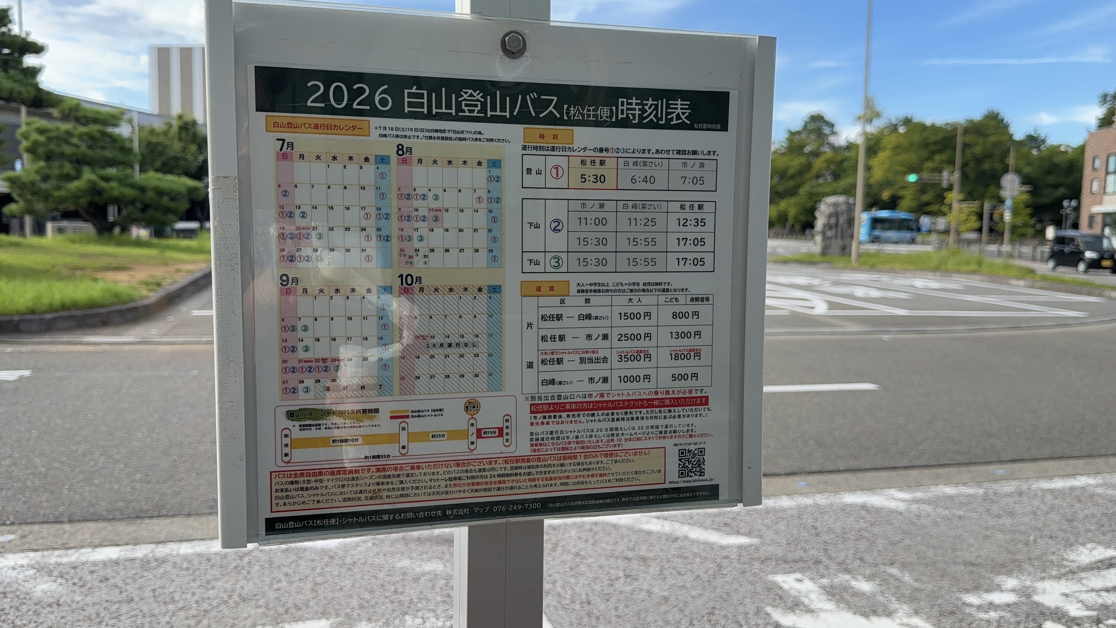 「2026白山登山バス【松任便】時刻表」の掲示板、駅前ロータリーを背景に