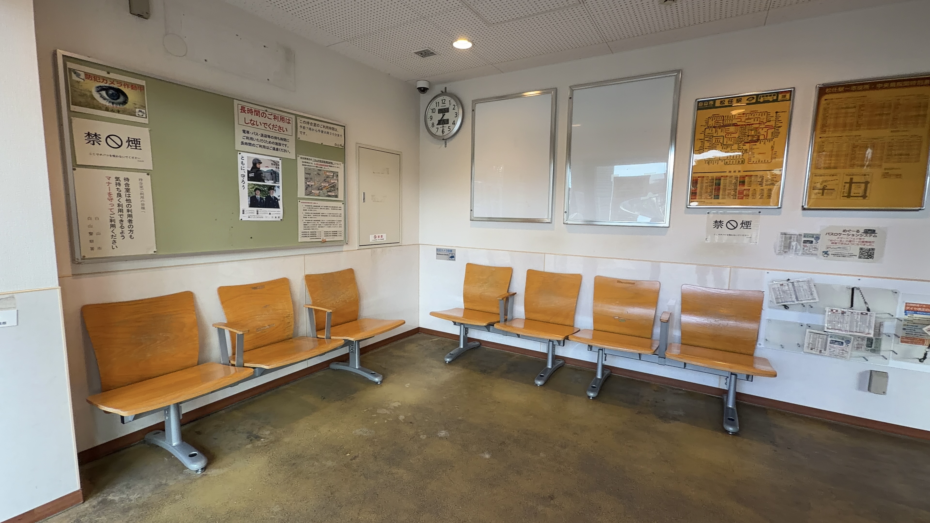 駅の待合室、木製のベンチが並び、時刻表や地図の掲示物、壁の時計