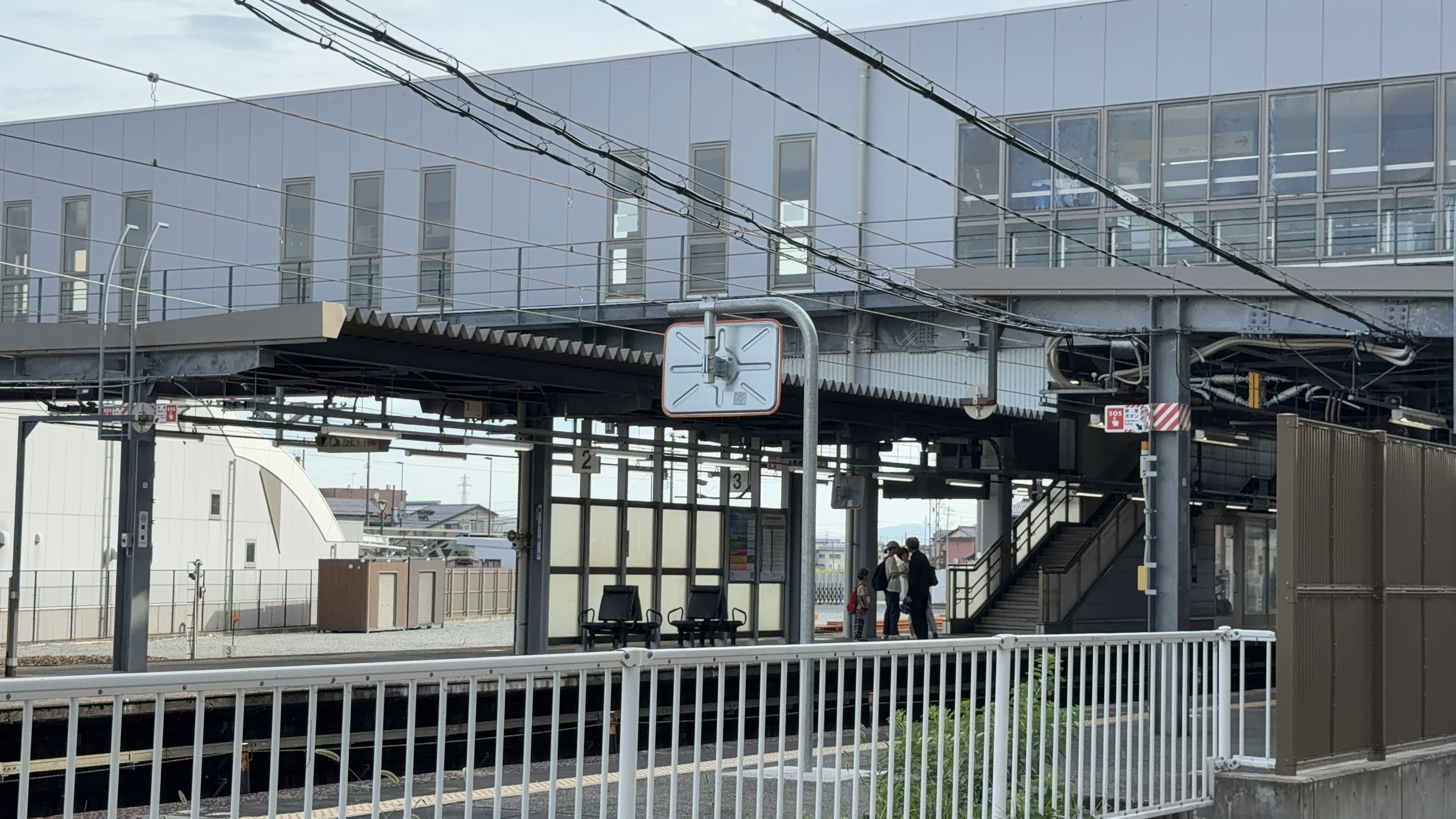 白い柵の階段越しに見える駅のホーム、跨線橋と発車案内、待つ人たち