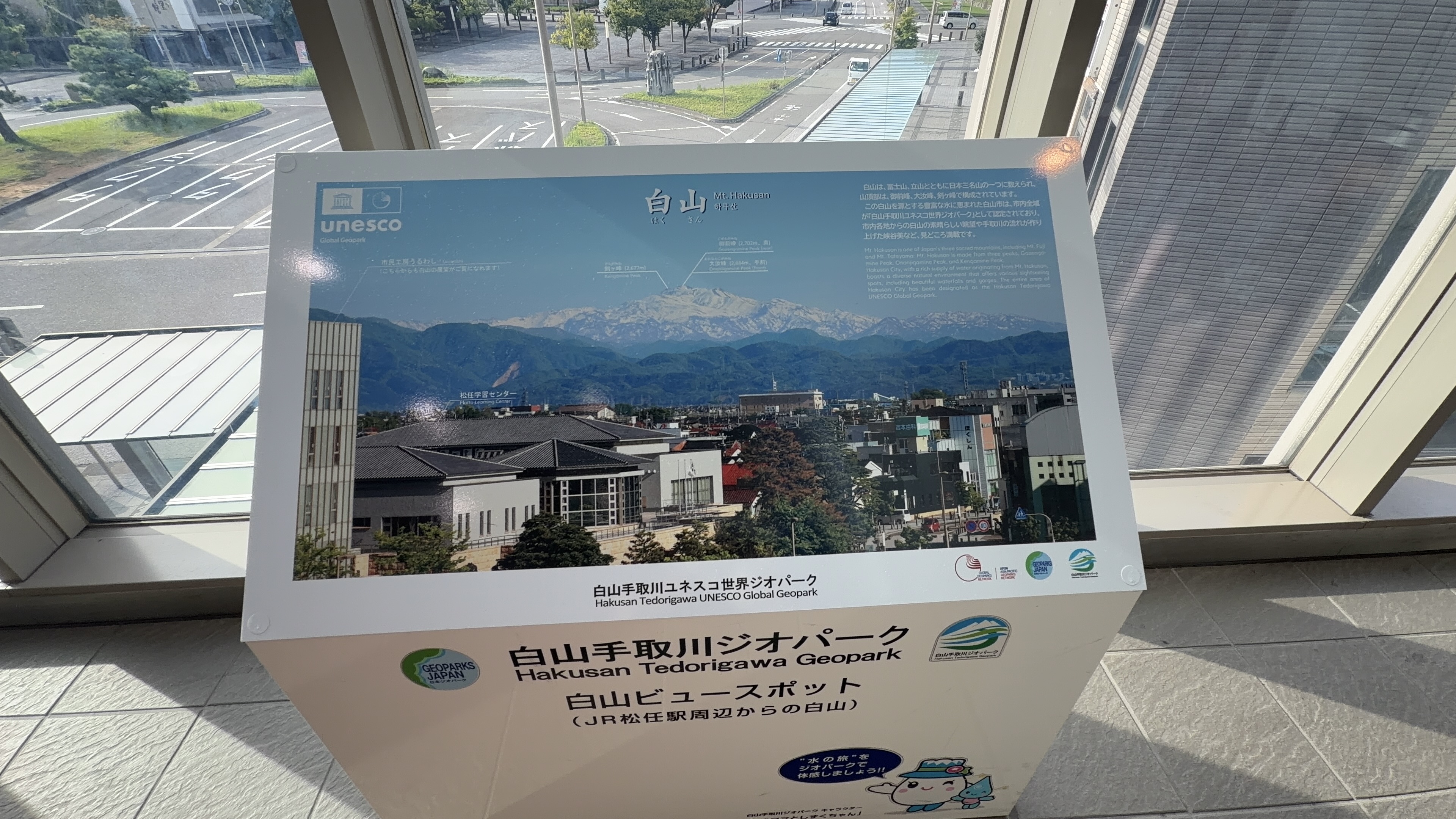 「白山手取川ジオパーク　白山ビュースポット」の案内板、白山の写真とユネスコのロゴ、窓の外に駅前ロータリー