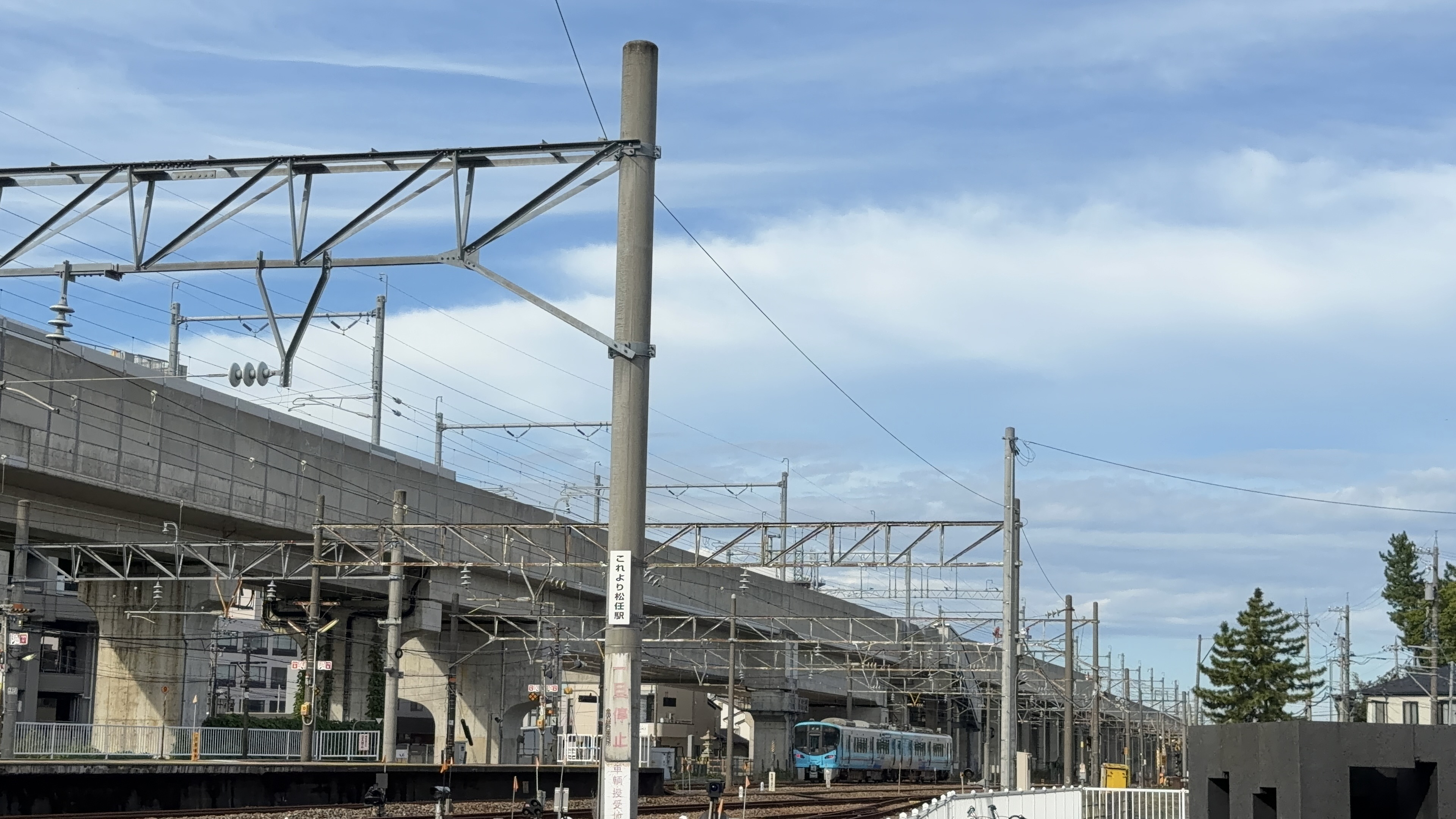 「これより松任駅」と書かれた標識のポール、奥に接近するIRいしかわ鉄道の青い車両と高架橋