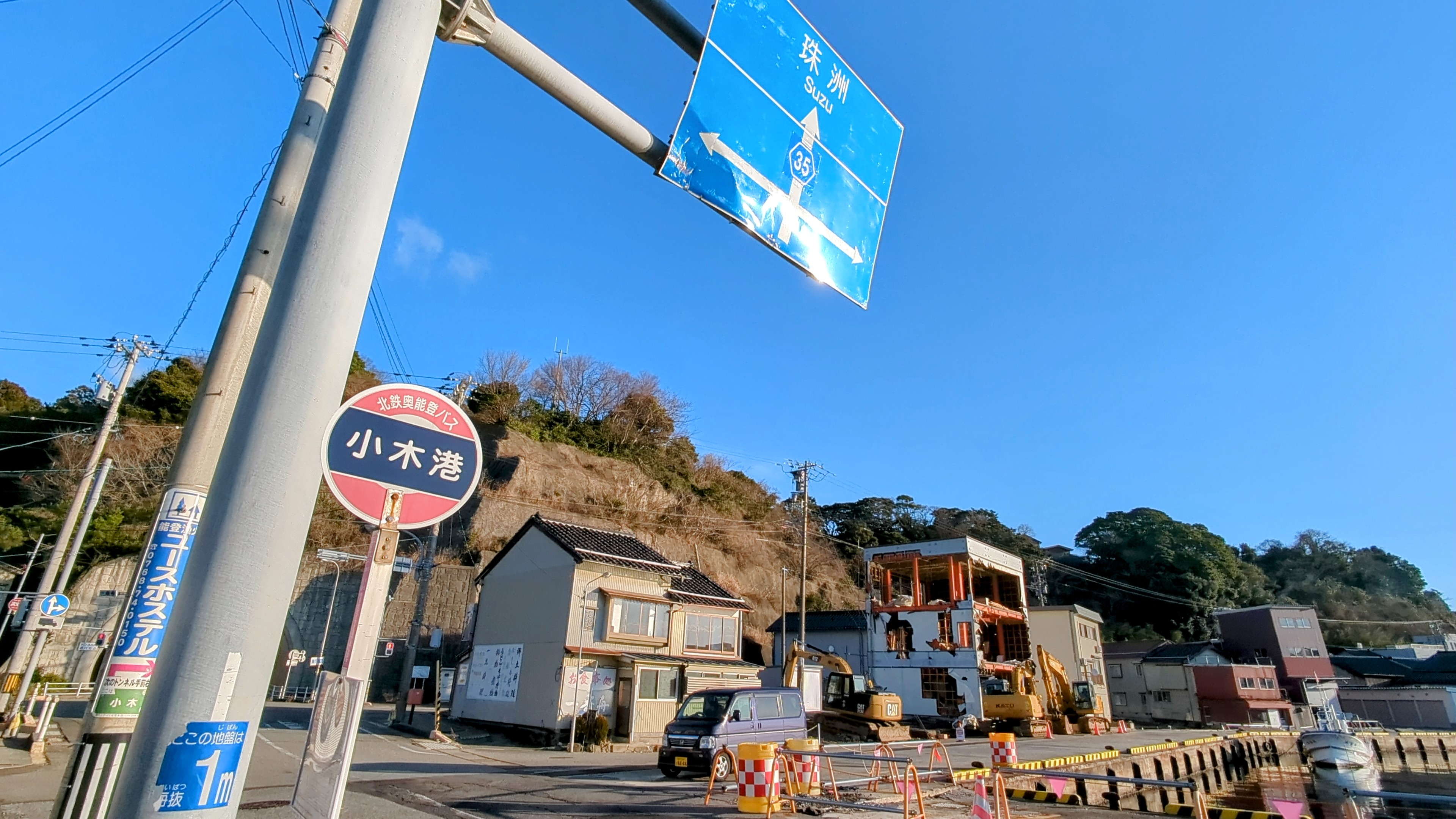 「小木港」のバス停標識、奥で重機が解体工事をしている様子、2025年1月
