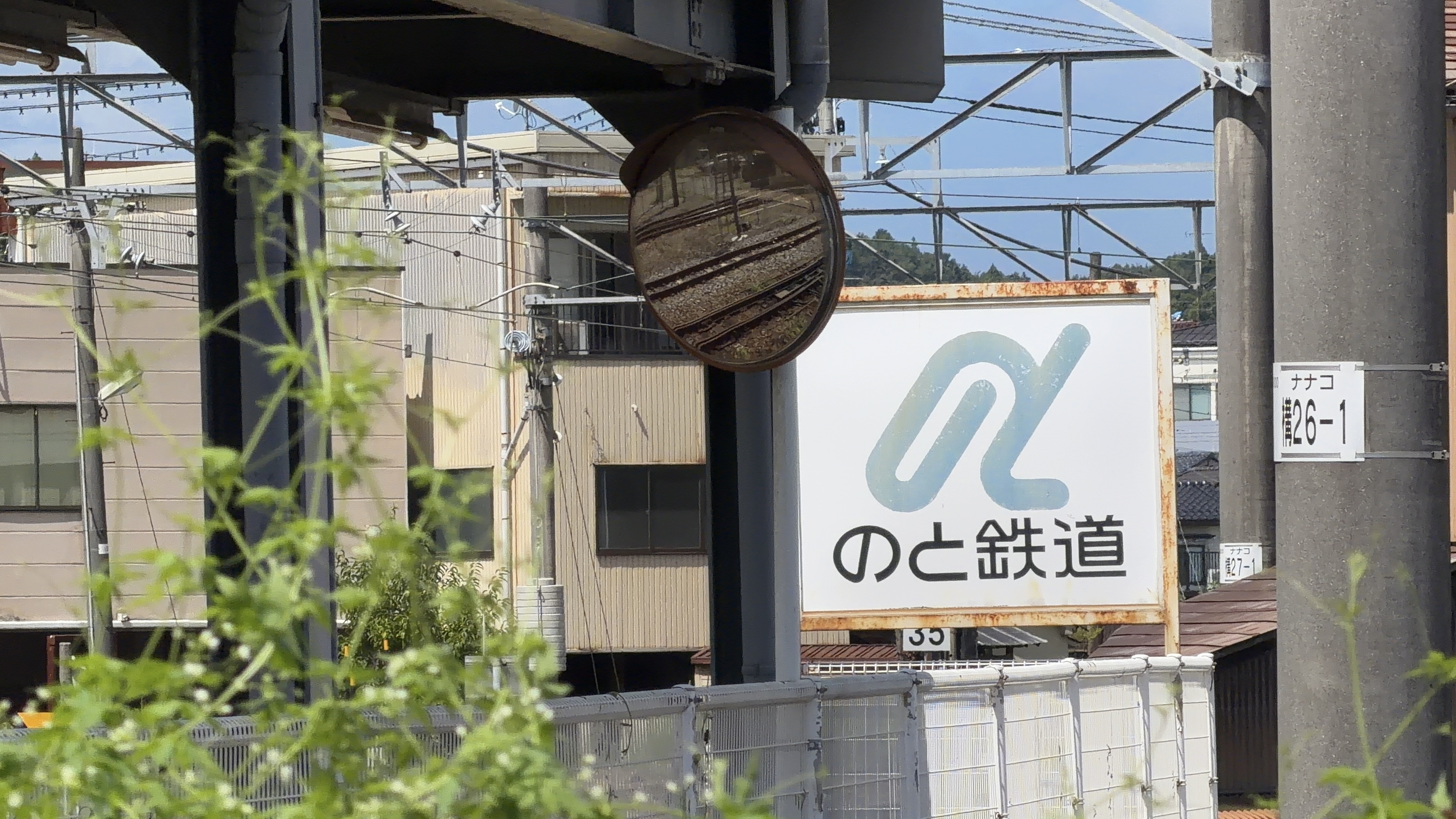 「のと鉄道」の看板、丸みを帯びたロゴマーク、奥に建ち並ぶ住宅と山なみ