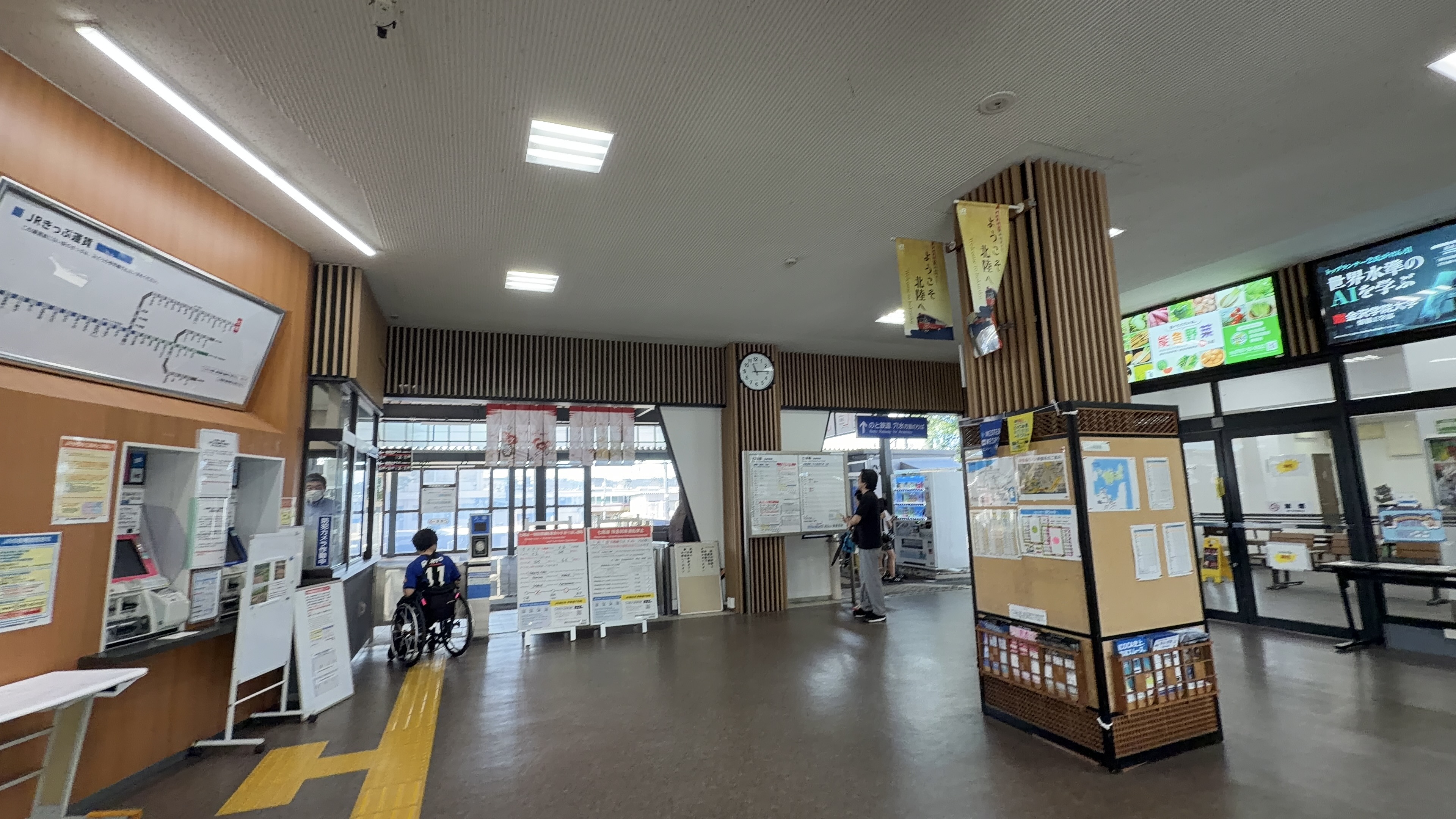 JRの鉄道案内所の窓口がある駅舎内、車椅子の方が窓口を待っている様子、天井から「ようこそ北陸へ」の旗
