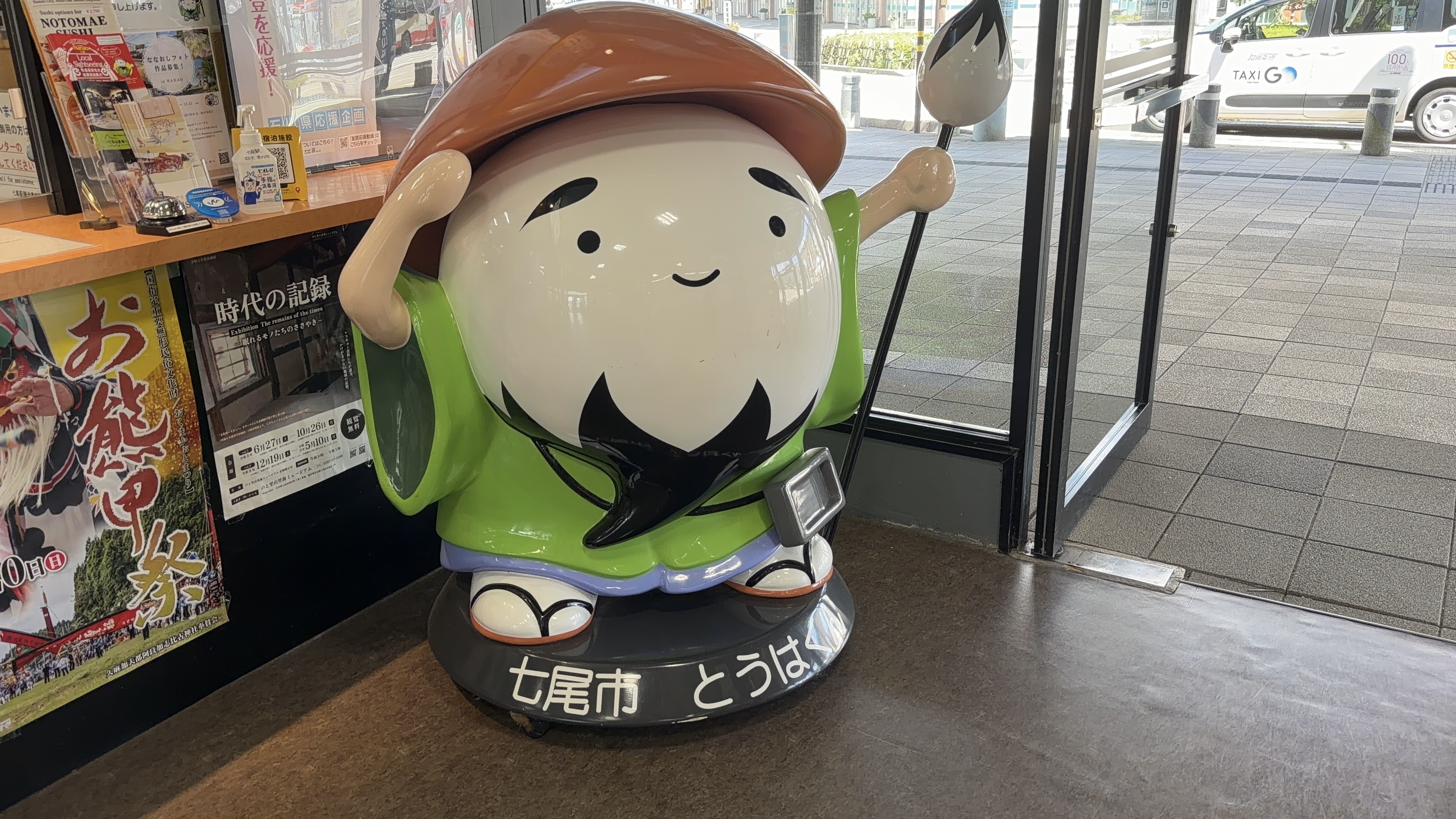 七尾市のマスコットキャラクター「とうはく」の立像、緑の着物姿で駅舎の出入口に立つ