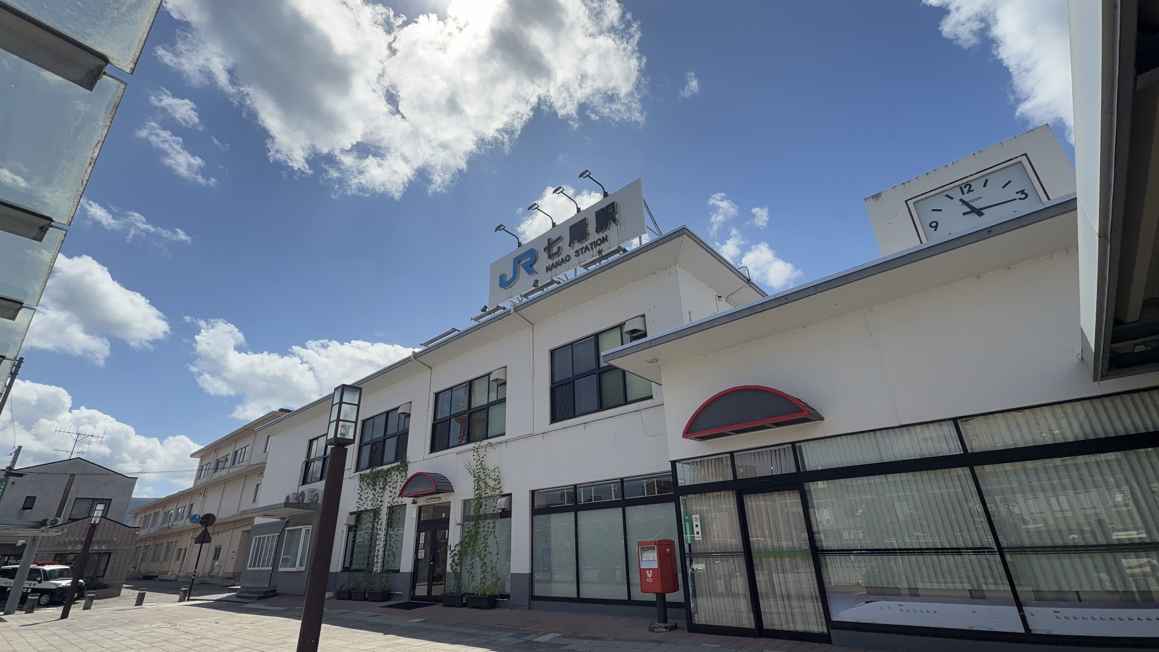 JR七尾駅の駅舎外観、屋上に「JR七尾駅 NANAO STATION」の看板と丸い時計、奥に交番とパトカー