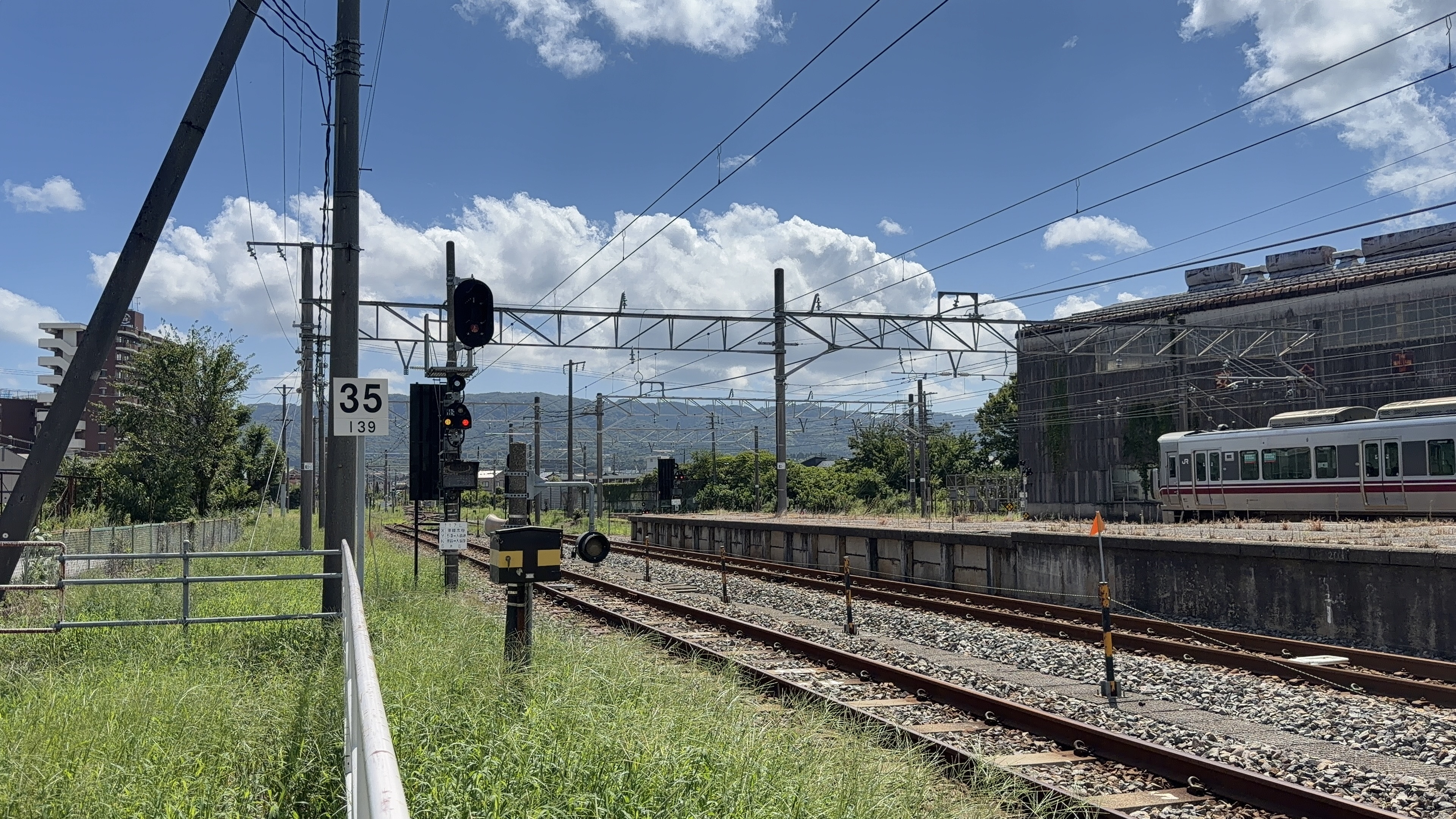 線路沿いの「35」の速度標識、信号機、線路の奥に車両基地と停まる電車、山なみ