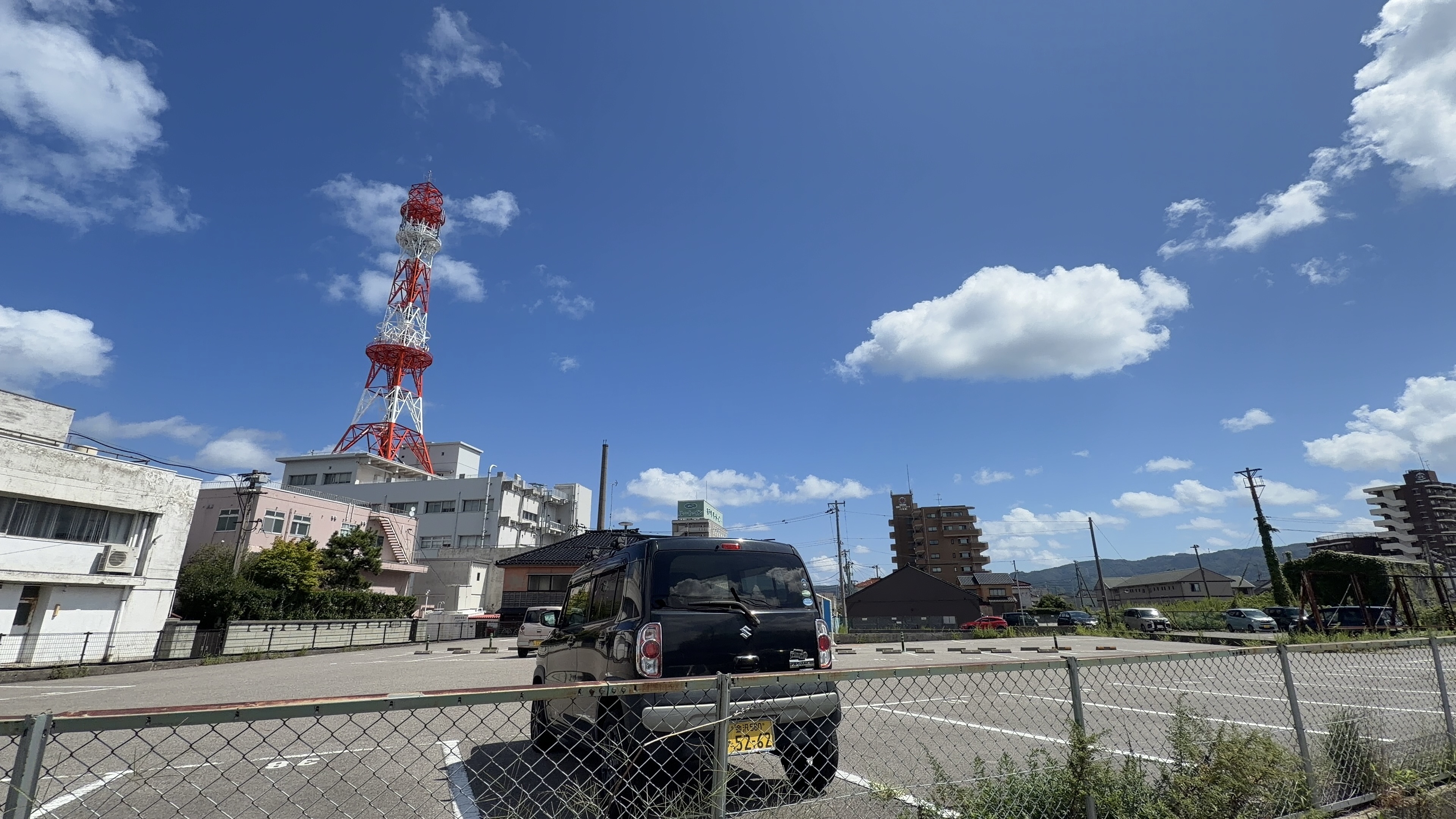 住宅街の駐車場、赤白の電波塔、黒い軽自動車が停まっている様子