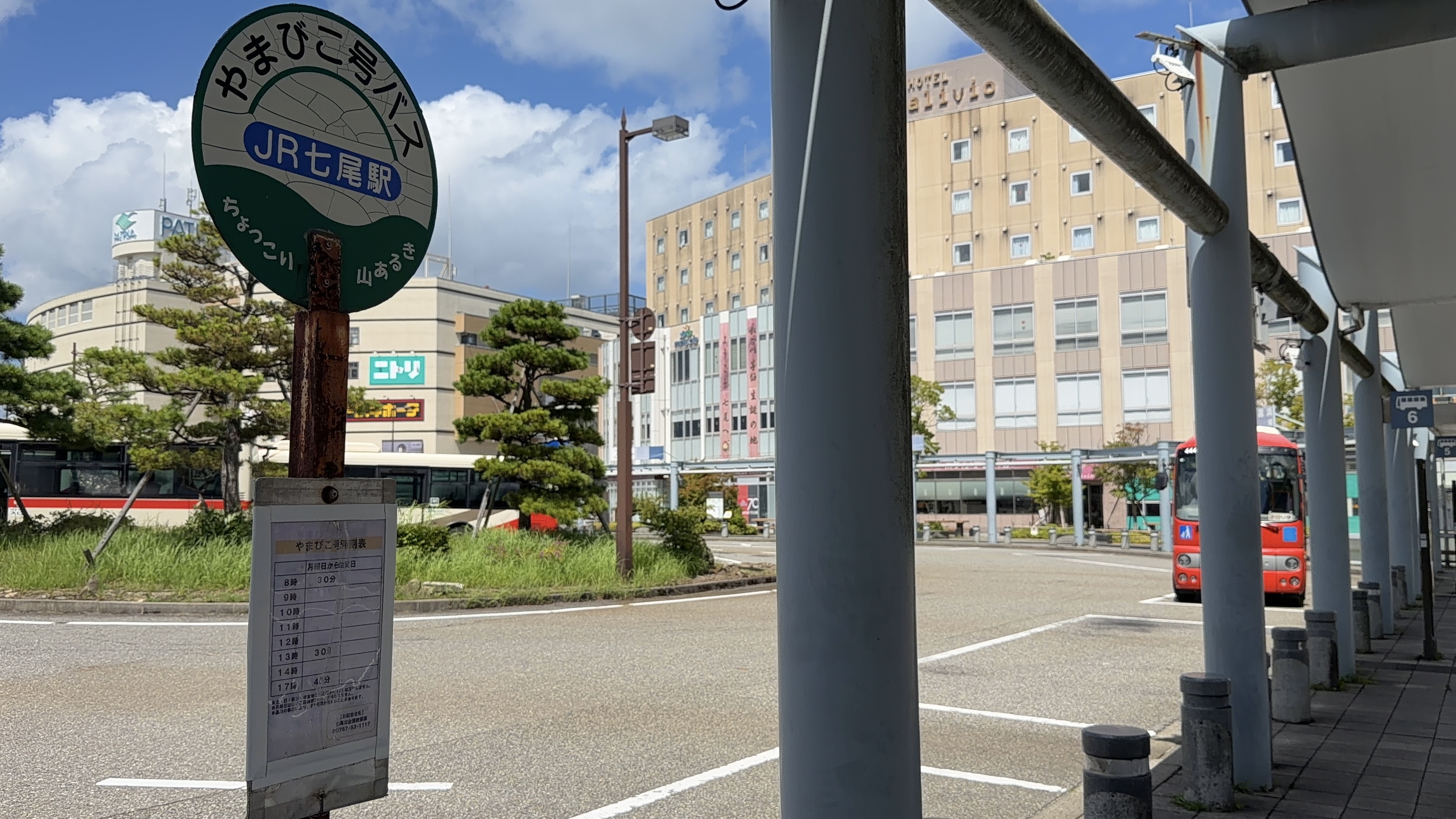 「やまびこ号バス JR七尾駅」のバス停ポール、時刻表、奥に赤いバスと「HOTEL alivio」の建物