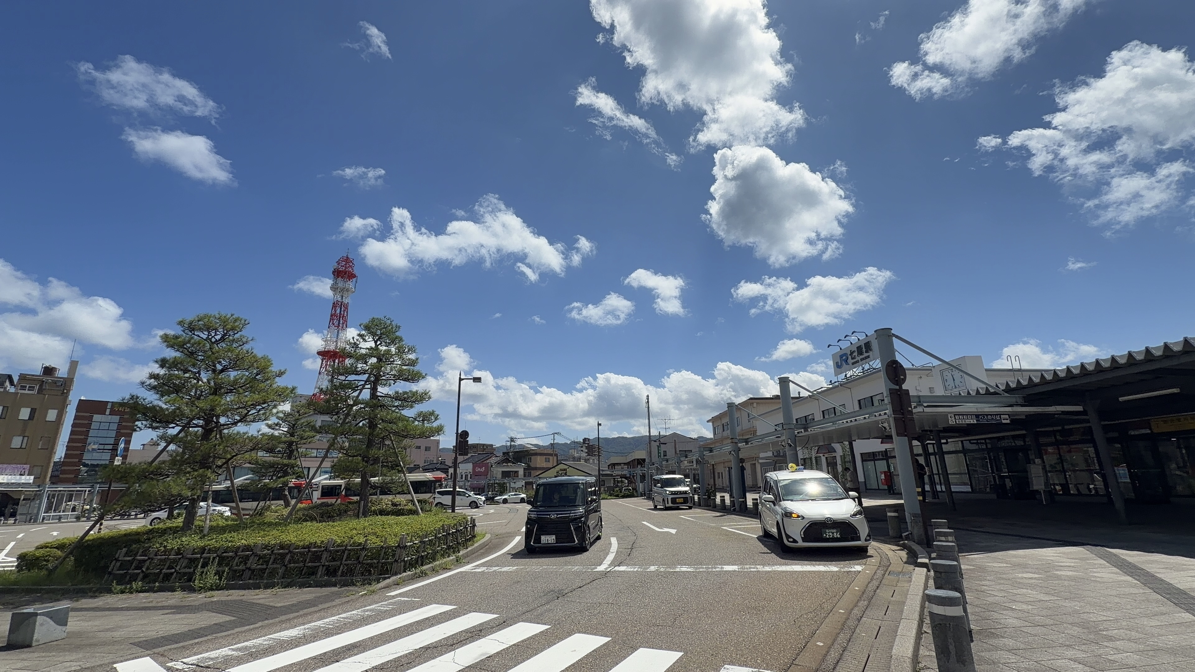 七尾駅前ロータリー、JR七尾駅の看板と赤白の電波塔、タクシーの並ぶ青空の下の駅前広場