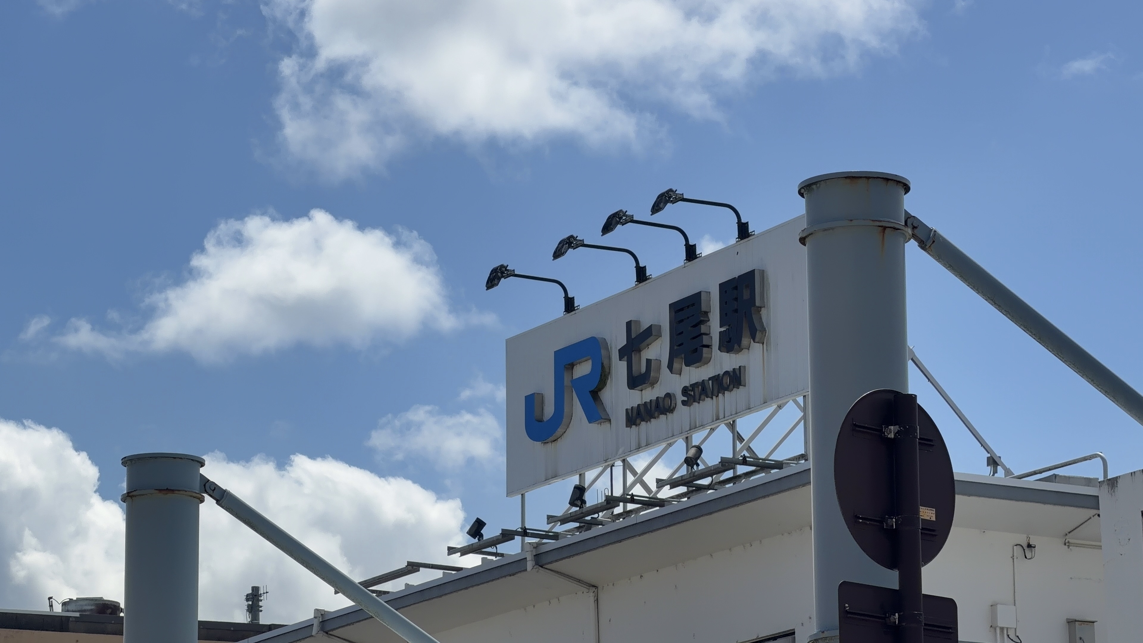 「JR七尾駅 NANAO STATION」の看板のクローズアップ、青いJRロゴと青空