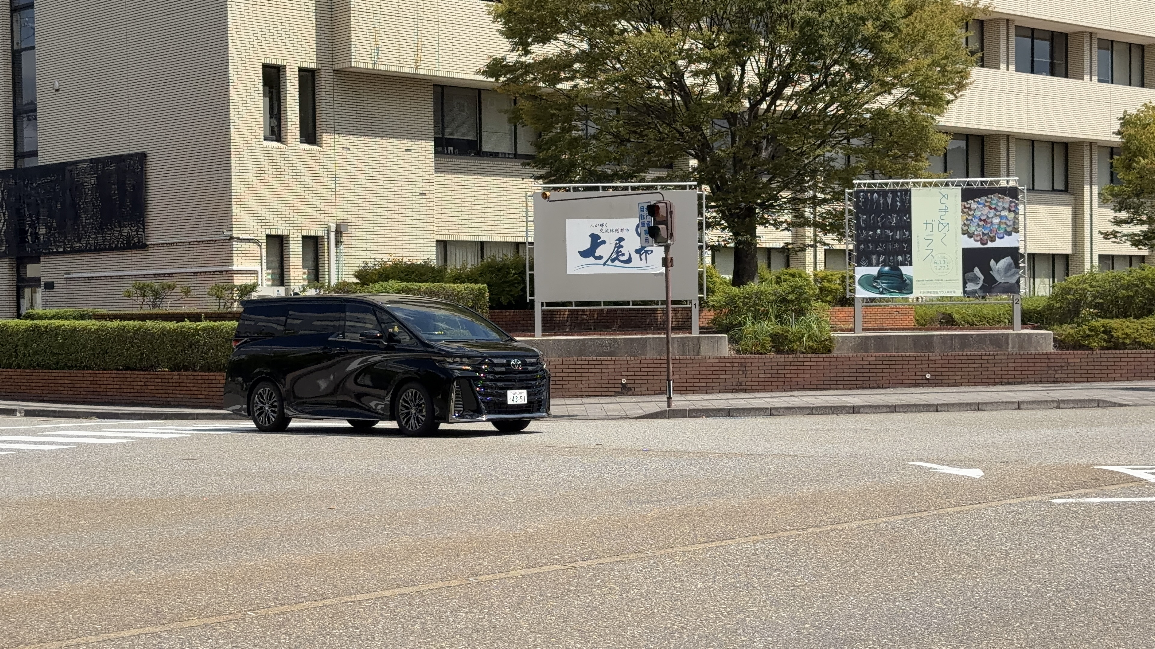 七尾市役所前の庭園、「人が輝く交流体感都市 七尾市」の看板前を通る黒いミニバン