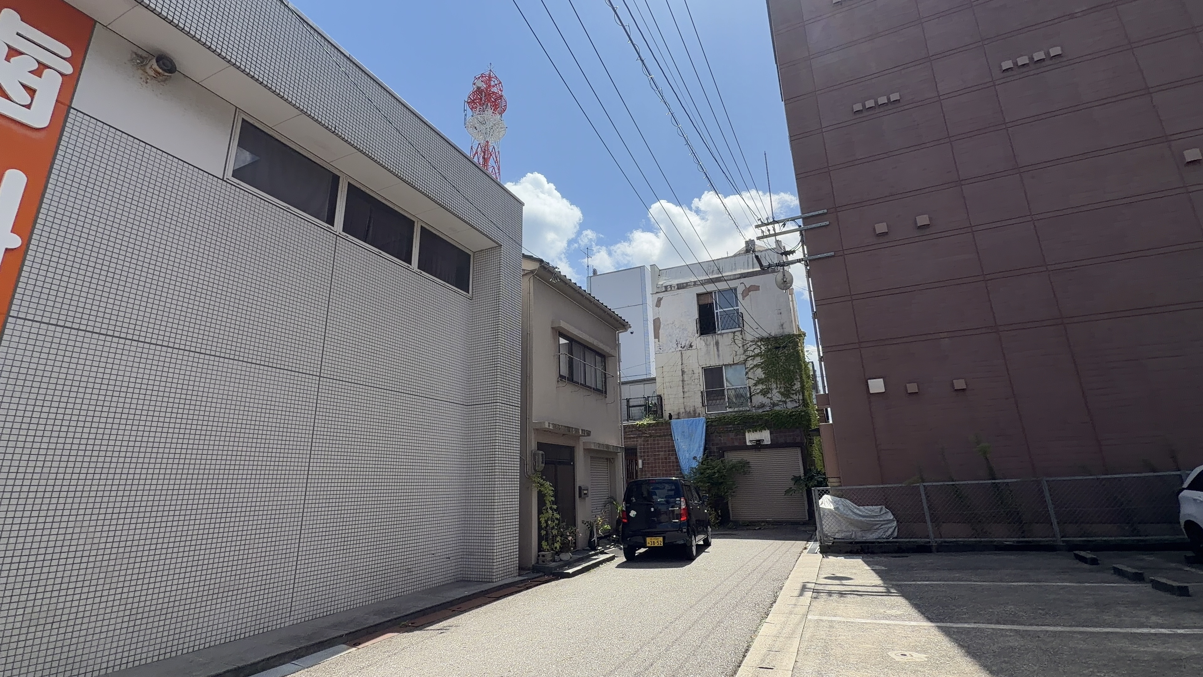 路地裏の細い通り、奥に赤白の電波塔、蔦の絡まる古い建物と黒い軽自動車