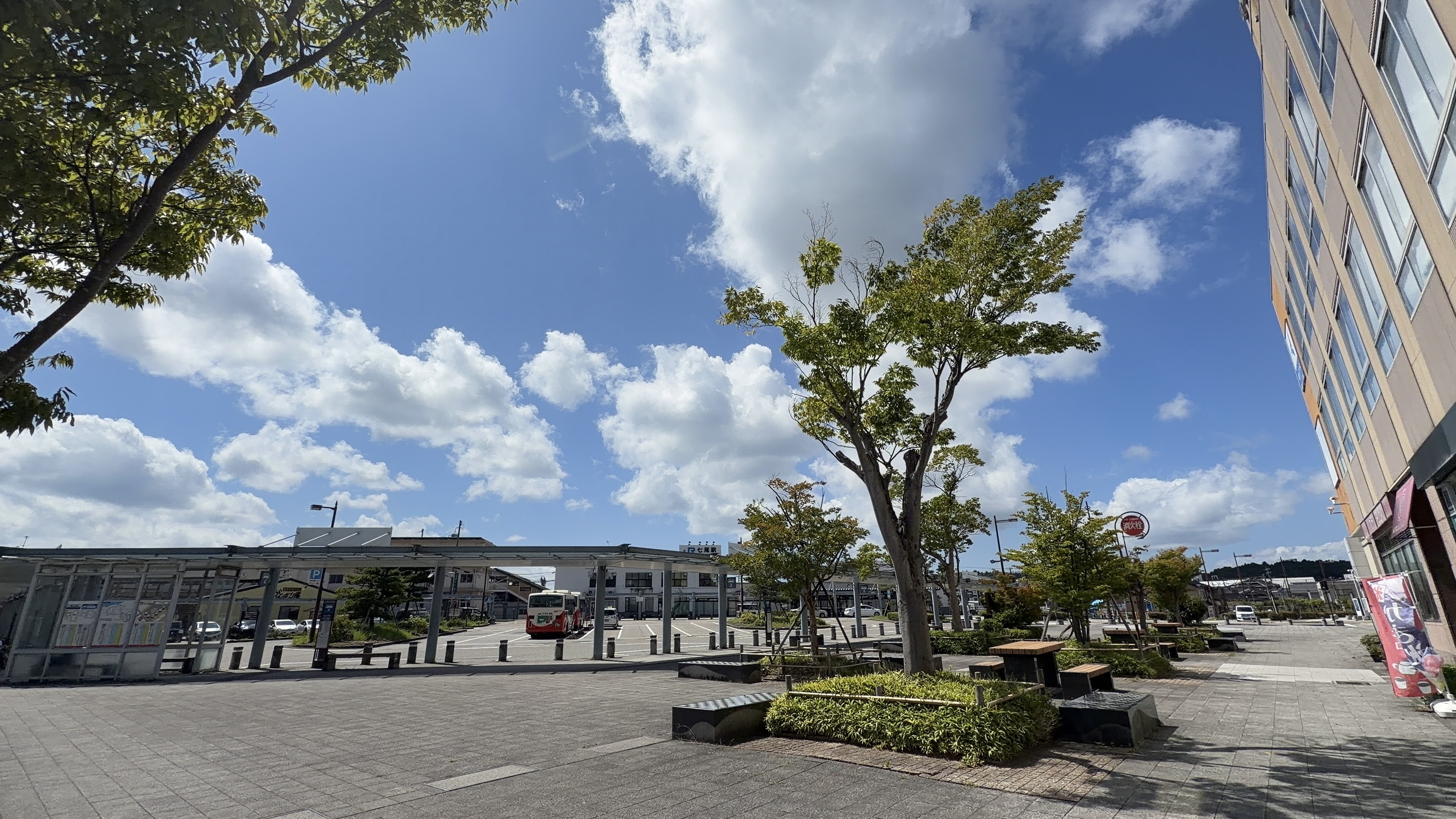七尾駅前広場、バスとベンチ、木立の向こうに駅舎が見える青空の広場
