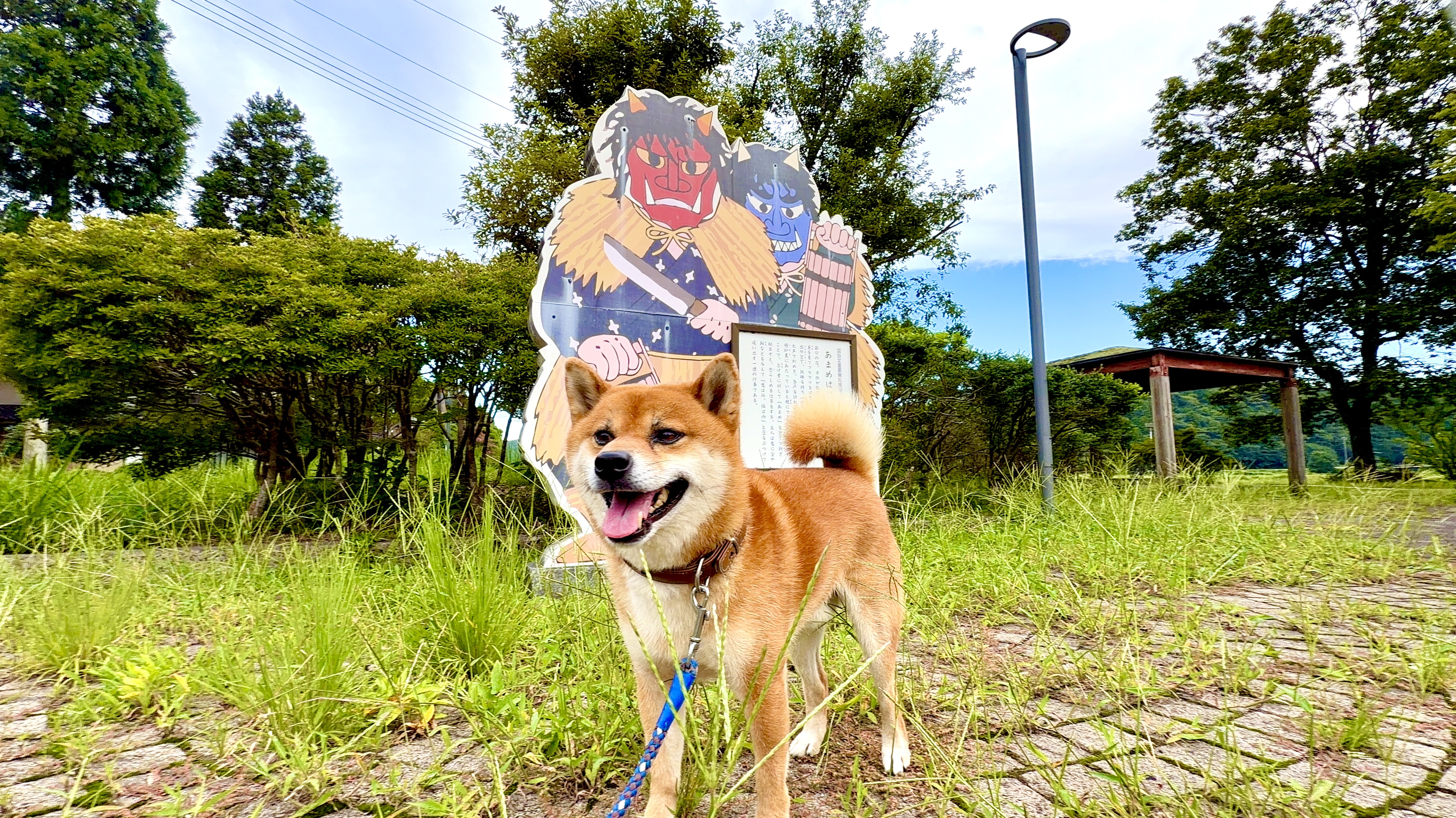 「あまめはぎ」の立看板の前で、しっぽを立てて笑顔のたろーさん。赤鬼と青鬼のイラストとともに