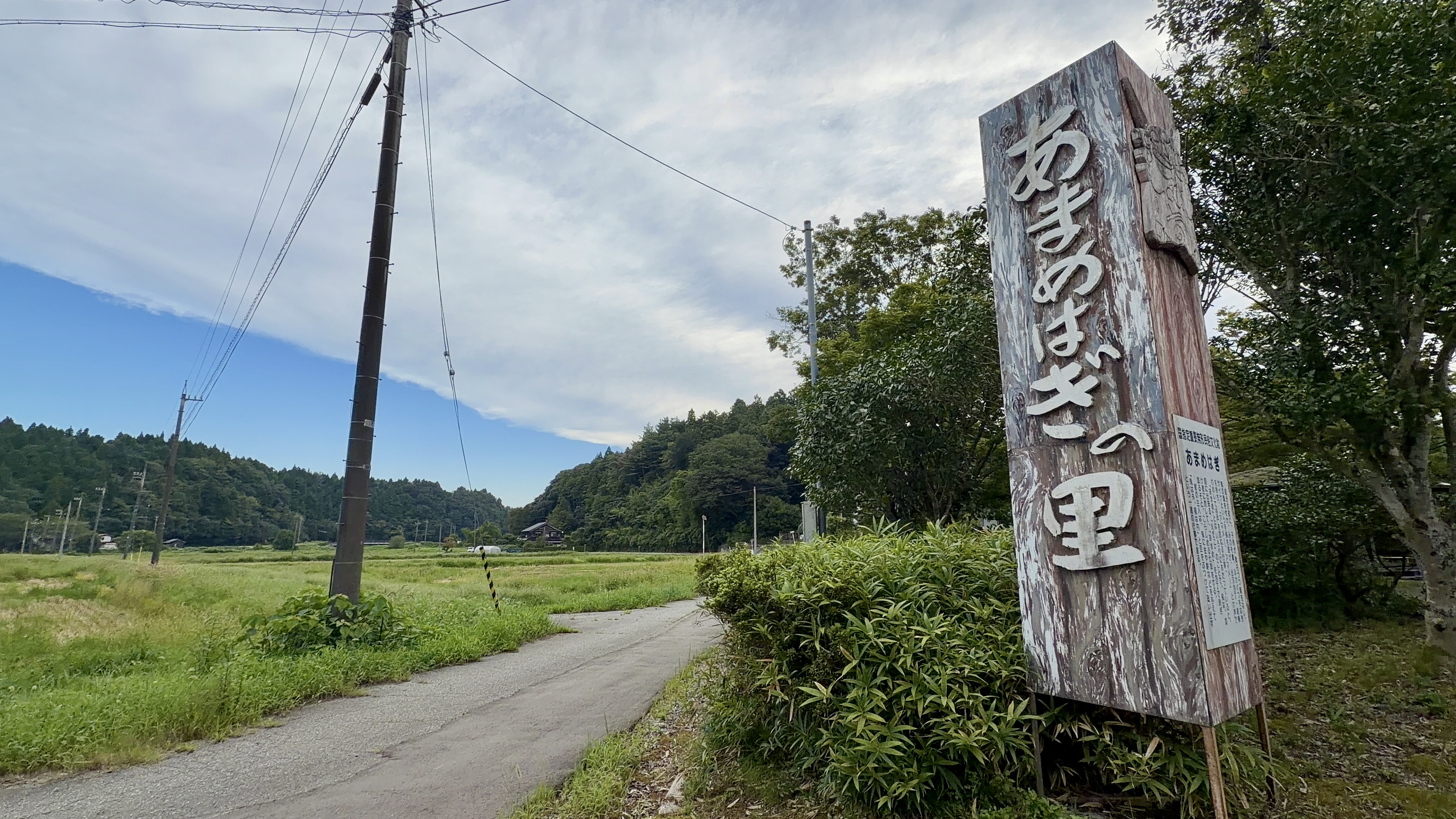 「あまめはぎの里」と刻まれた木製の案内塔、奥に田んぼと山並み