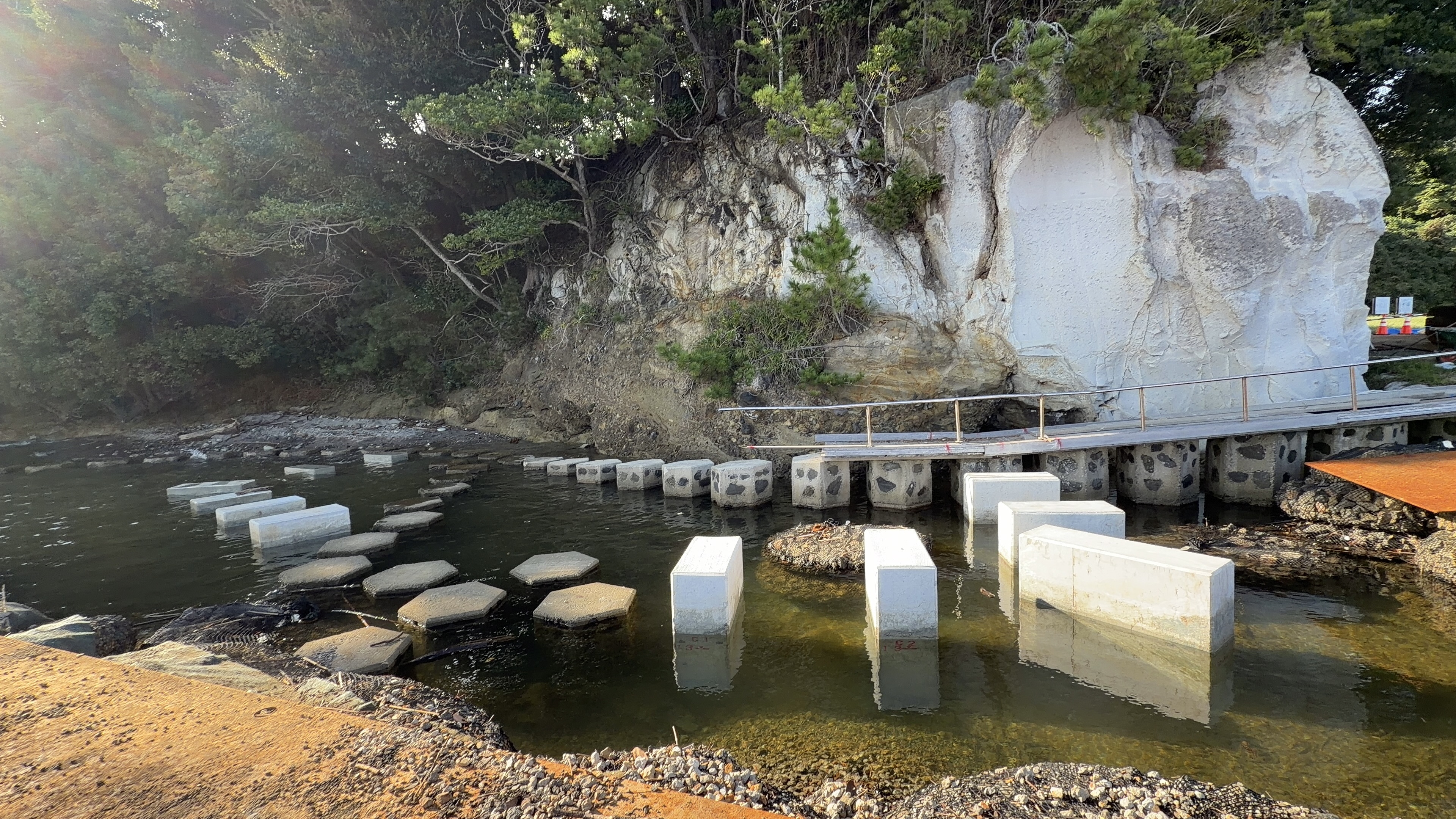 崖の下の浅瀬に並ぶ、白いコンクリートブロックと灰色の飛び石。奥に手すり付きの遊歩道と工事用の看板が見える
