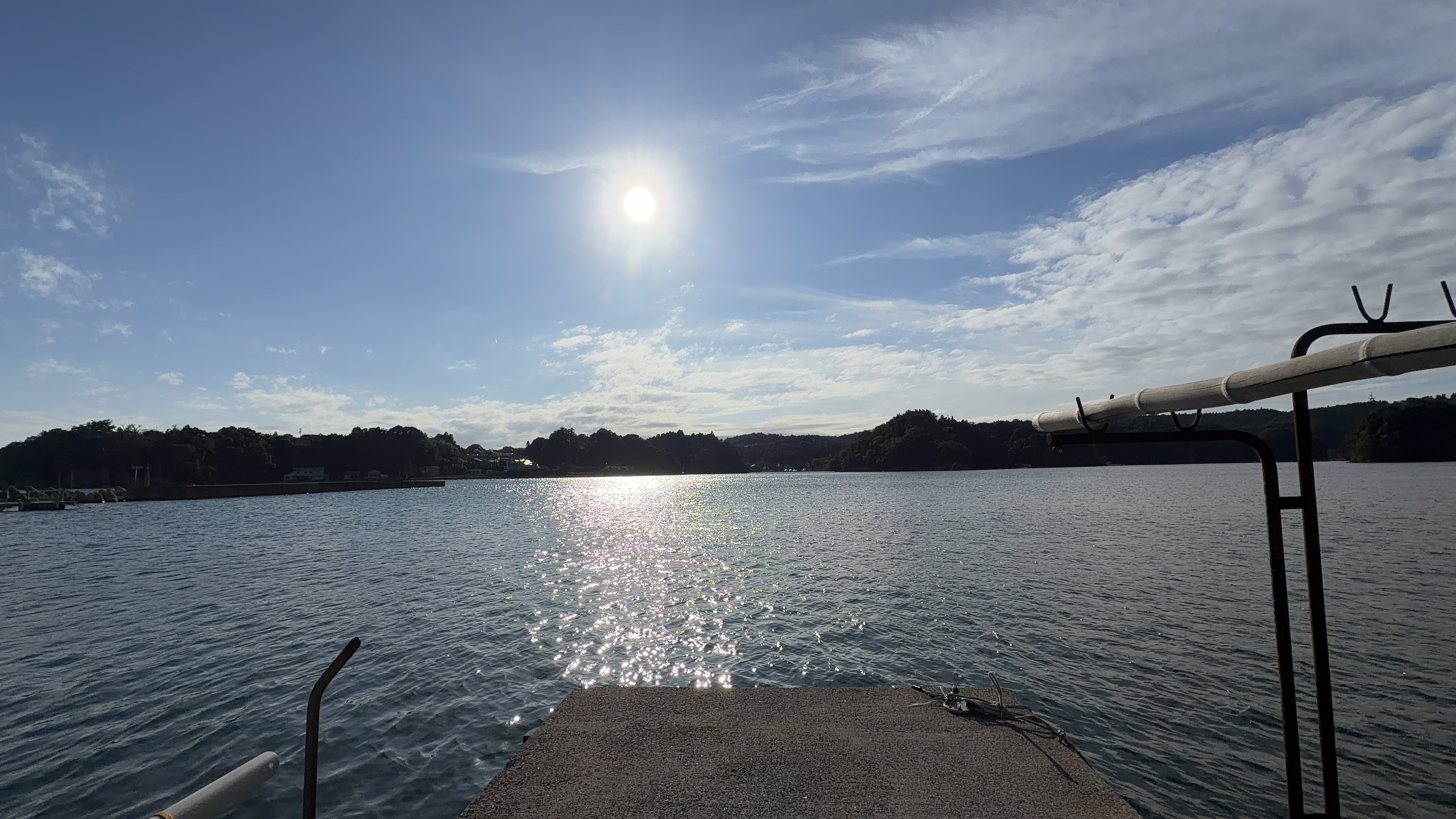 逆光の入り江、水面がきらきらと輝く。手前に釣り具の置かれた岸壁