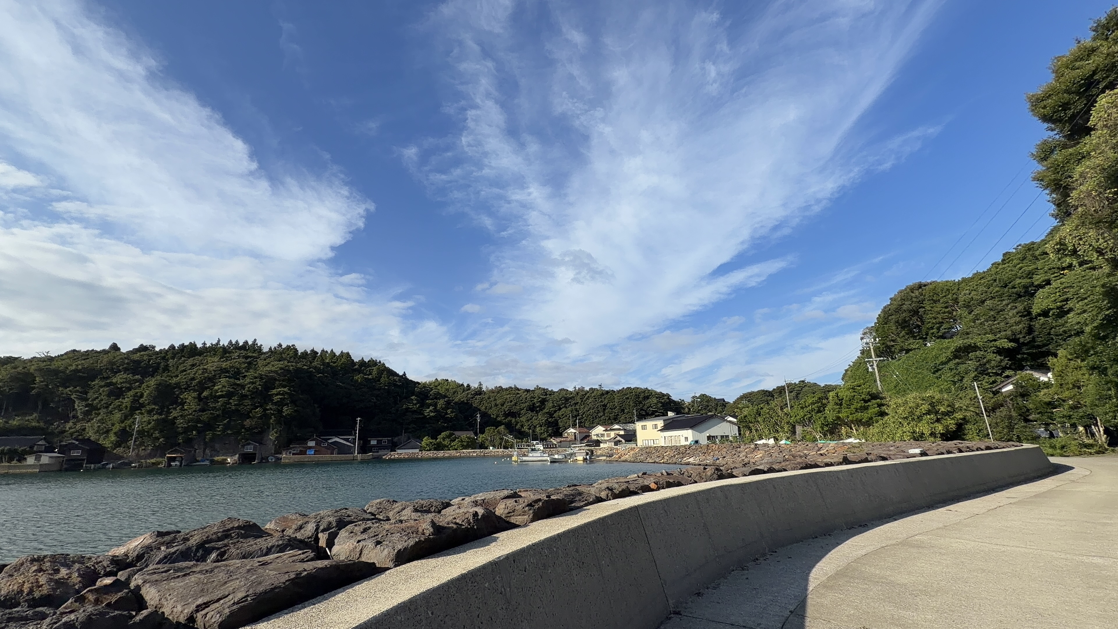 緩やかにカーブする護岸の道。左手に穏やかな入り江と漁村の家並み
