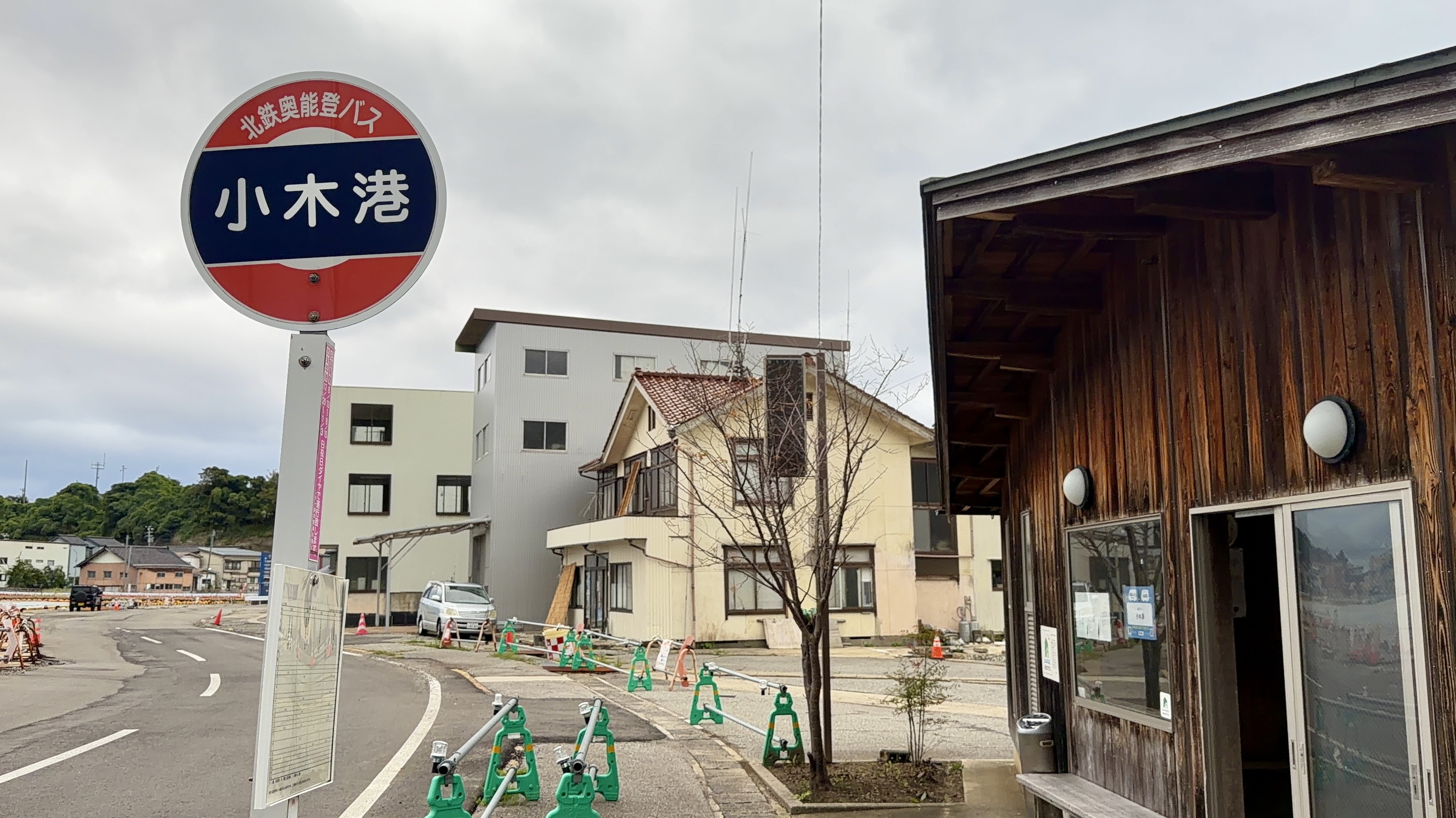 「小木港」バス停の看板と、奥に建ち並ぶ港沿いの建物