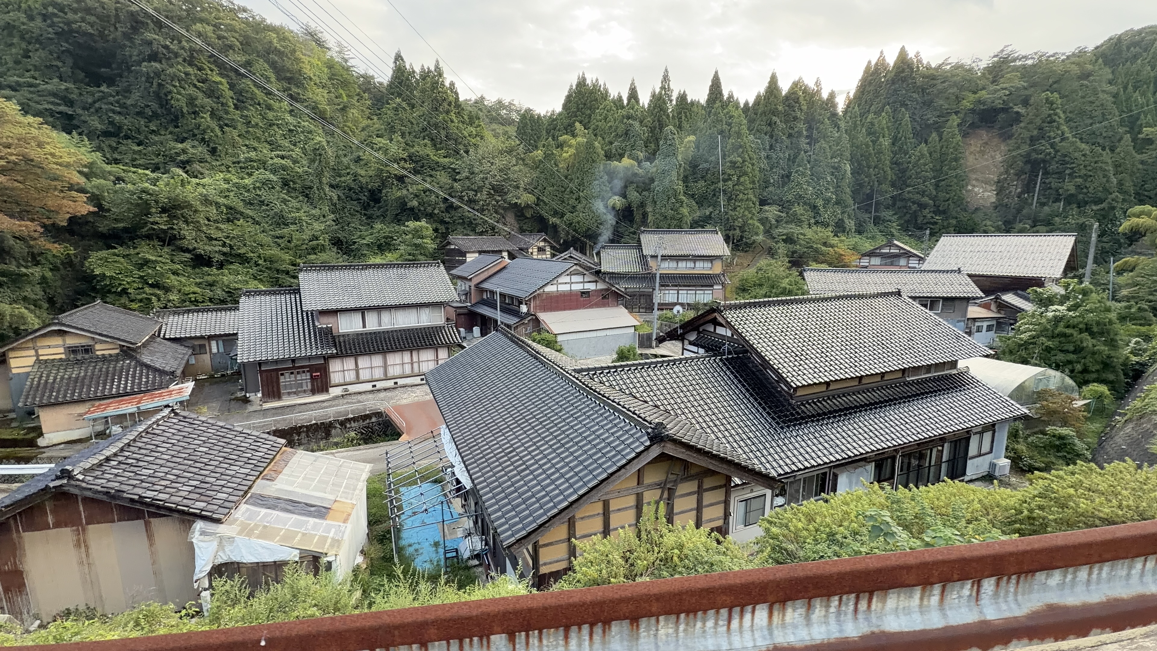 橋の上から見下ろす谷あいの集落、瓦屋根の家々が斜面に並び、奥に杉林の山