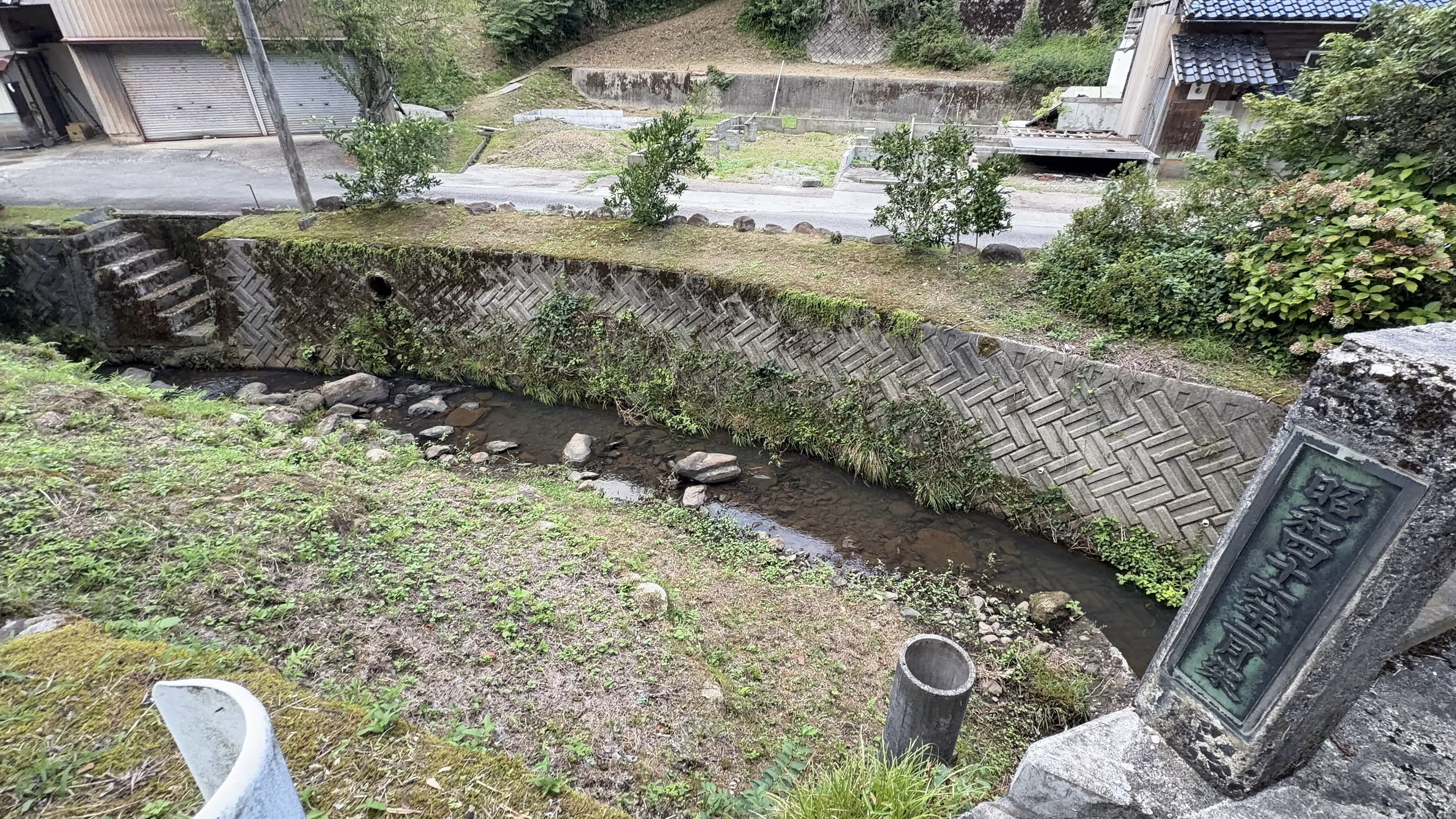 石積みの護岸に囲まれた小川、奥に空き地と工事中の建物、右手に古い町名標石