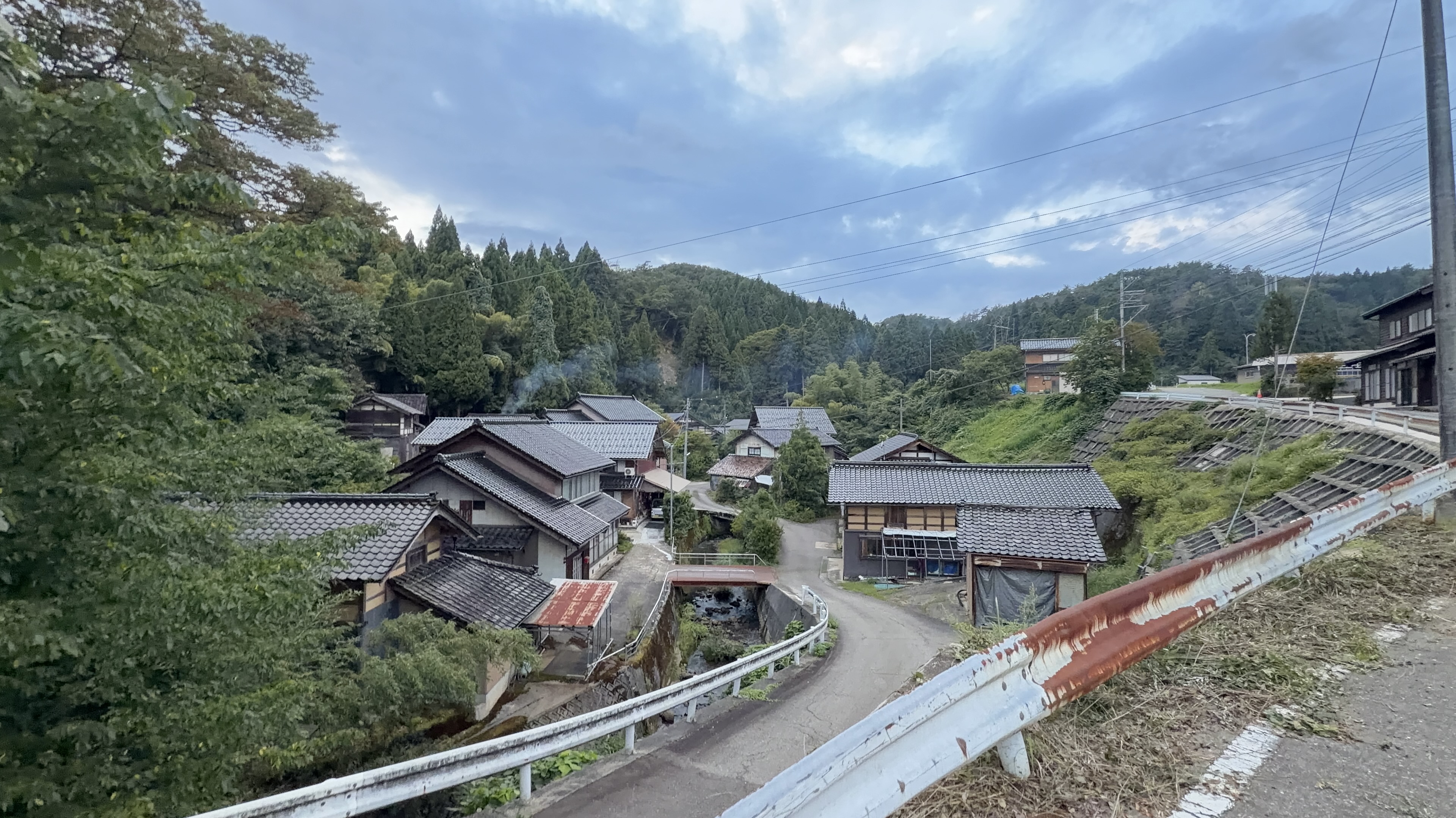 高台から見下ろす山あいの集落、瓦屋根の家々の間に赤い欄干の橋が見える、夕暮れの空