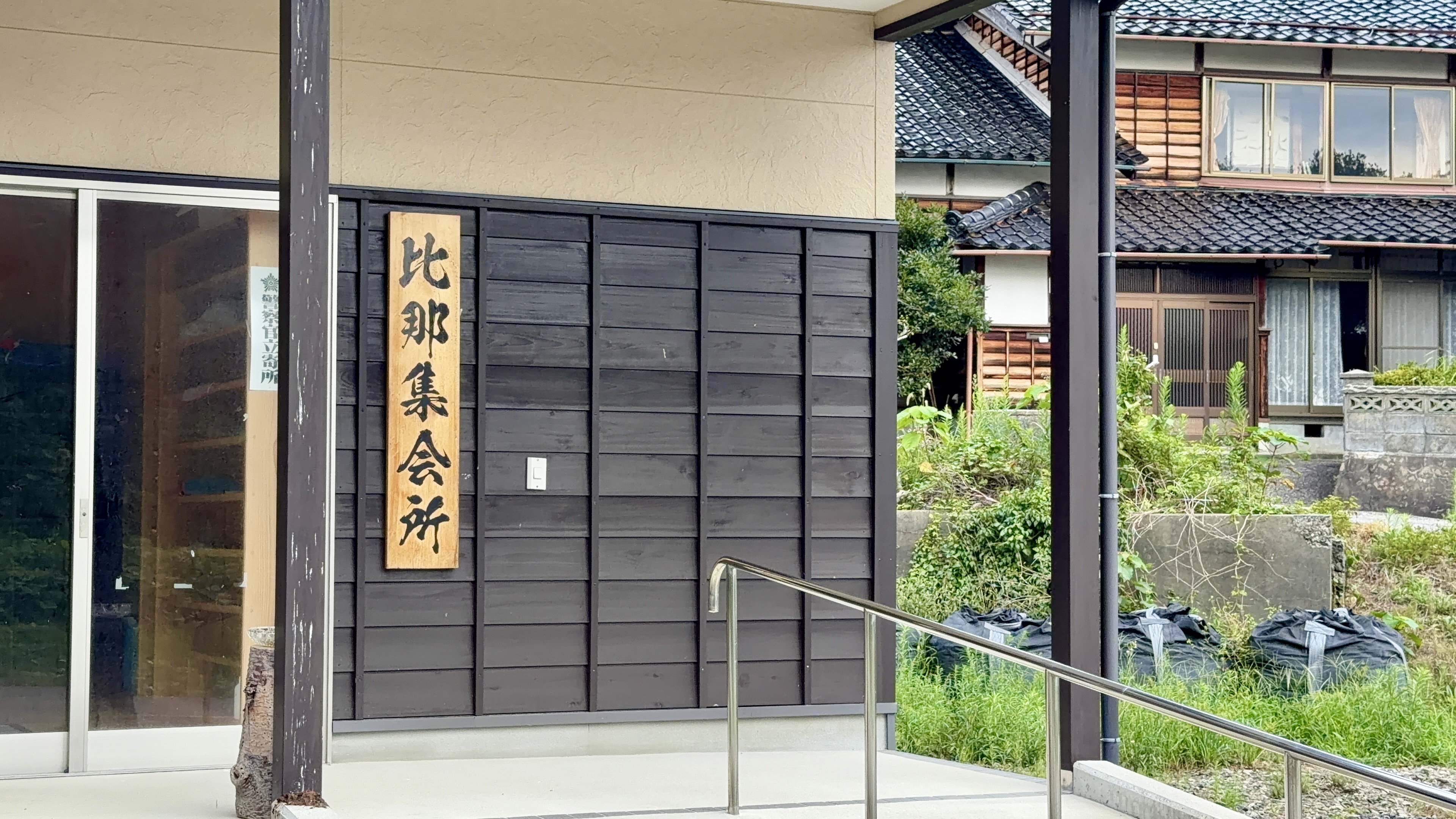 「比那集会所」の木の看板がかかる、暗い色の板壁の建物。奥に集落の家並みが見える