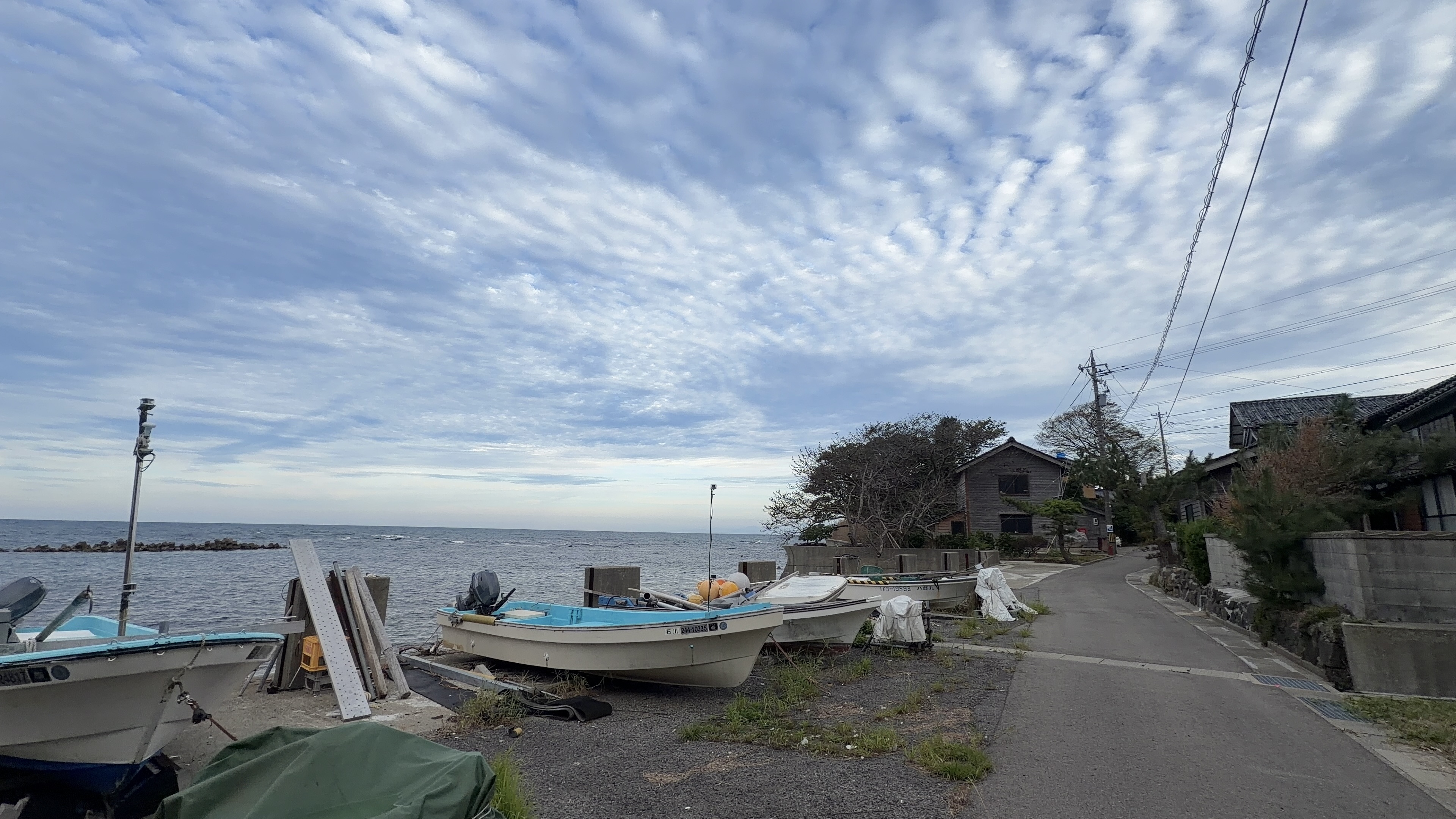 浜に上げられた漁船が並ぶ道、奥に電柱と家並み、うろこ雲の夕方の空