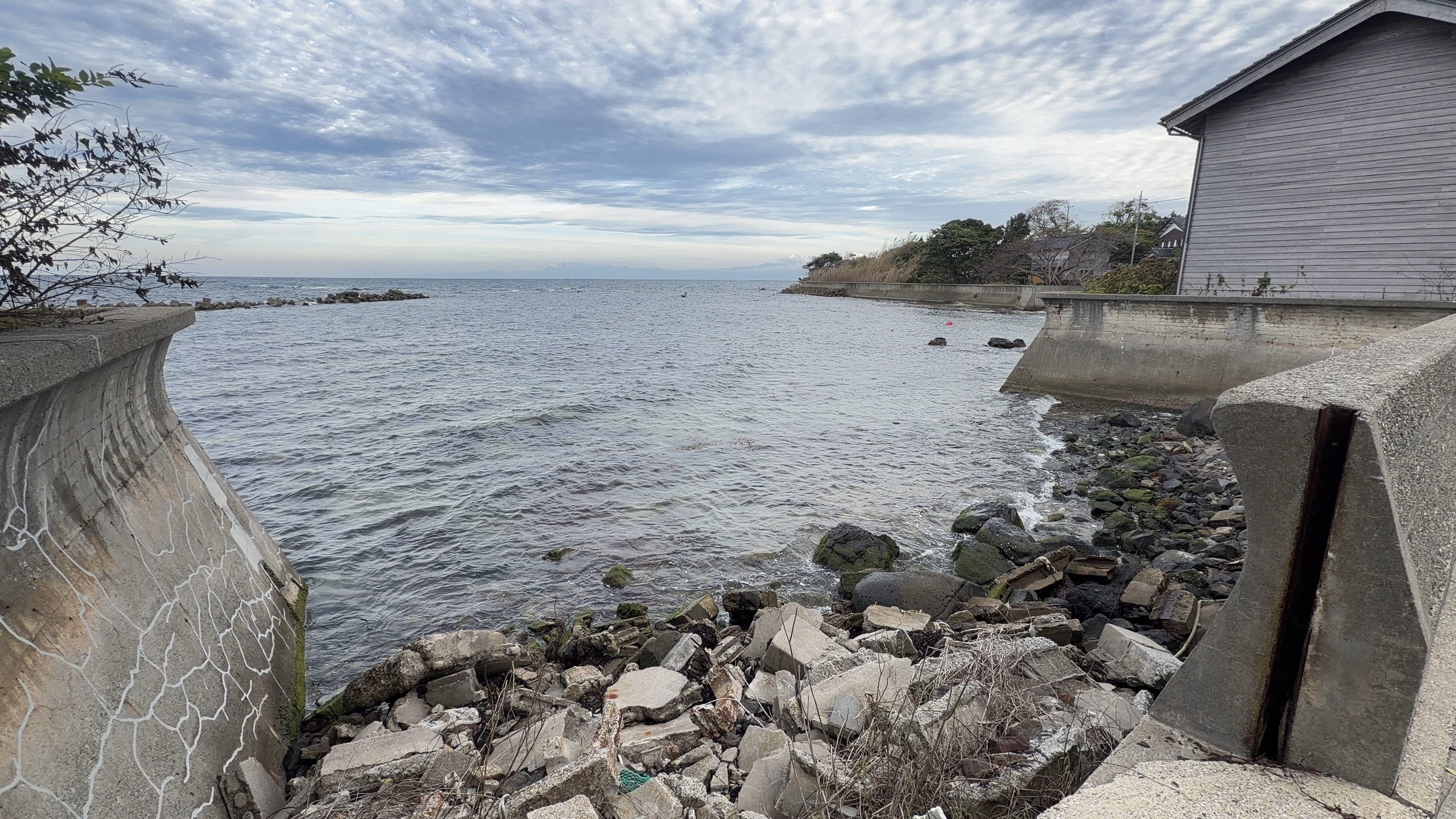 ひび割れたコンクリート護岸と、崩れたブロックが積み重なる海岸、奥に木造家屋