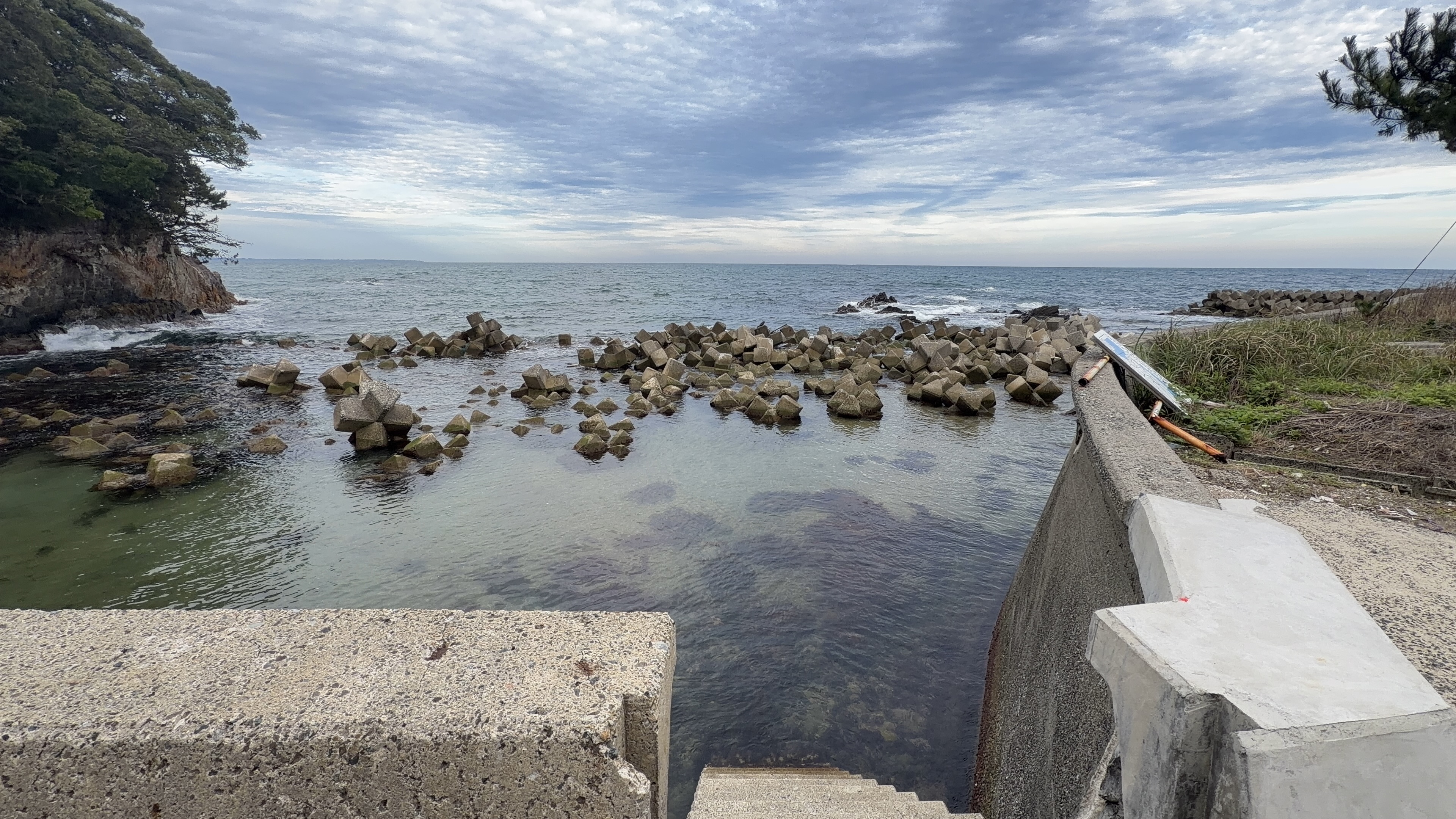 消波ブロックが並ぶ小さな入江、岬の緑と曇り空、コンクリート護岸の先の海