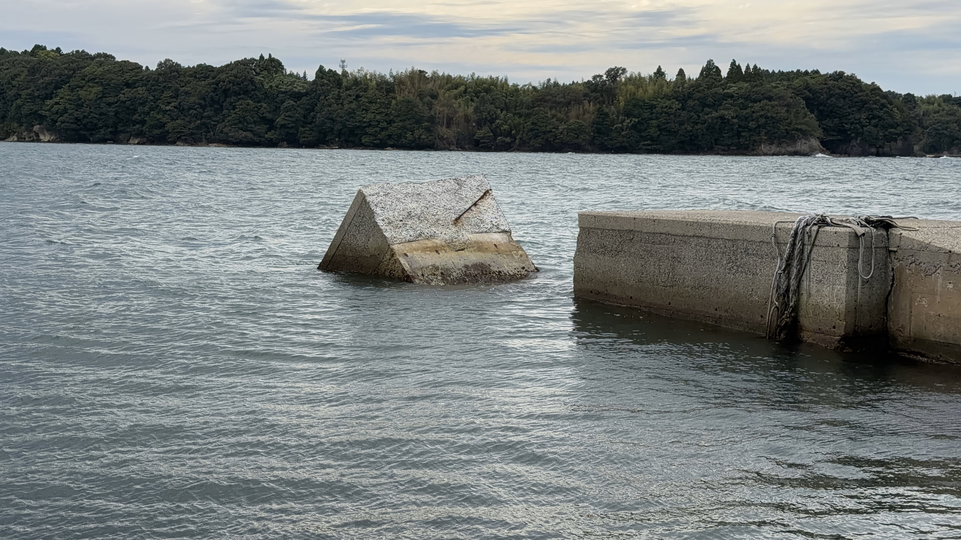 海に沈みかけた三角形のコンクリート構造物と、隣の防波堤、奥に緑の岬