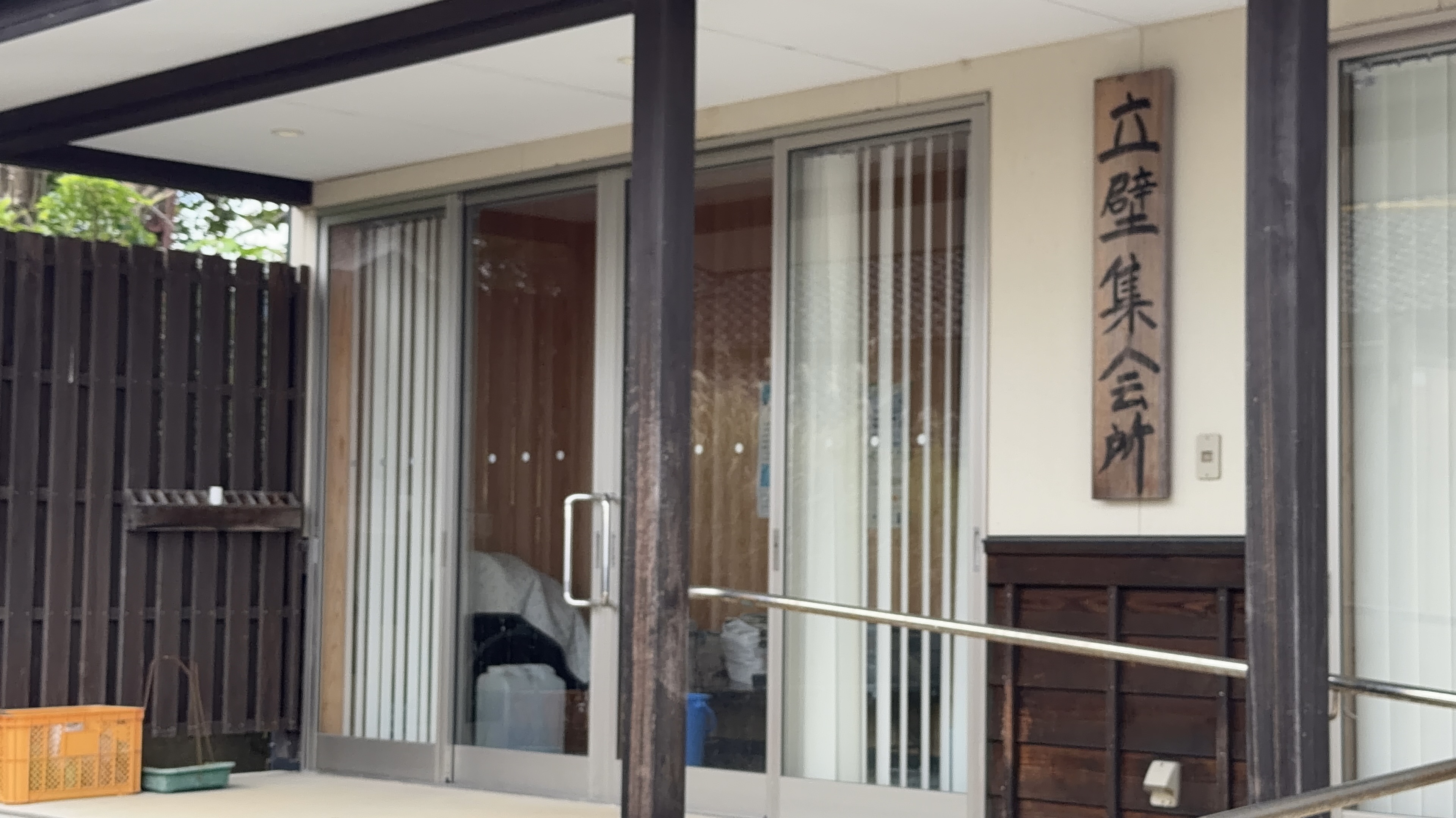 「立壁集会所」と書かれた木製看板、玄関の引き戸とオレンジ色のコンテナ