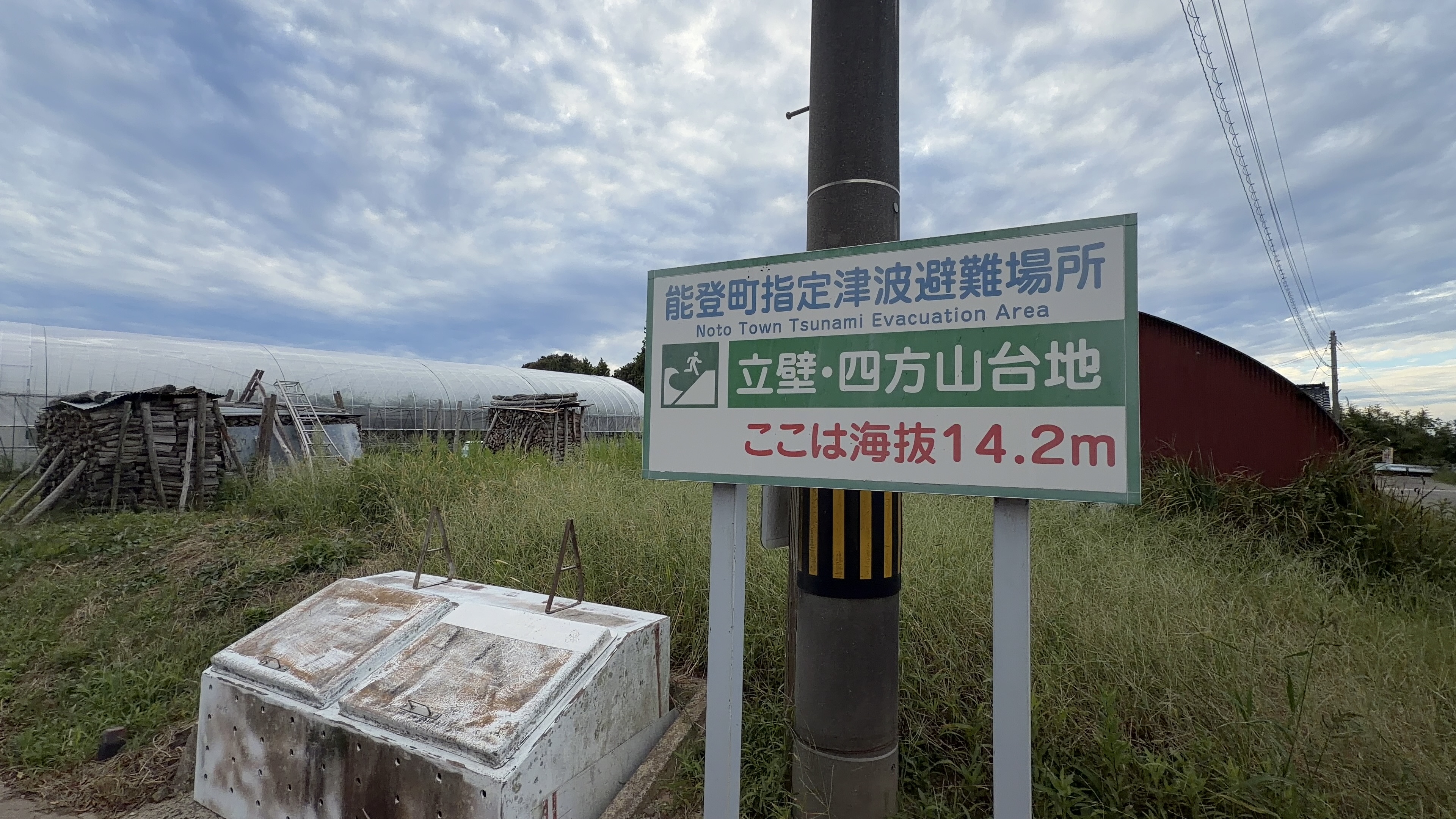 「能登町指定津波避難場所 立壁・四方山台地 ここは海抜14.2m」と書かれた案内看板、奥にビニールハウス